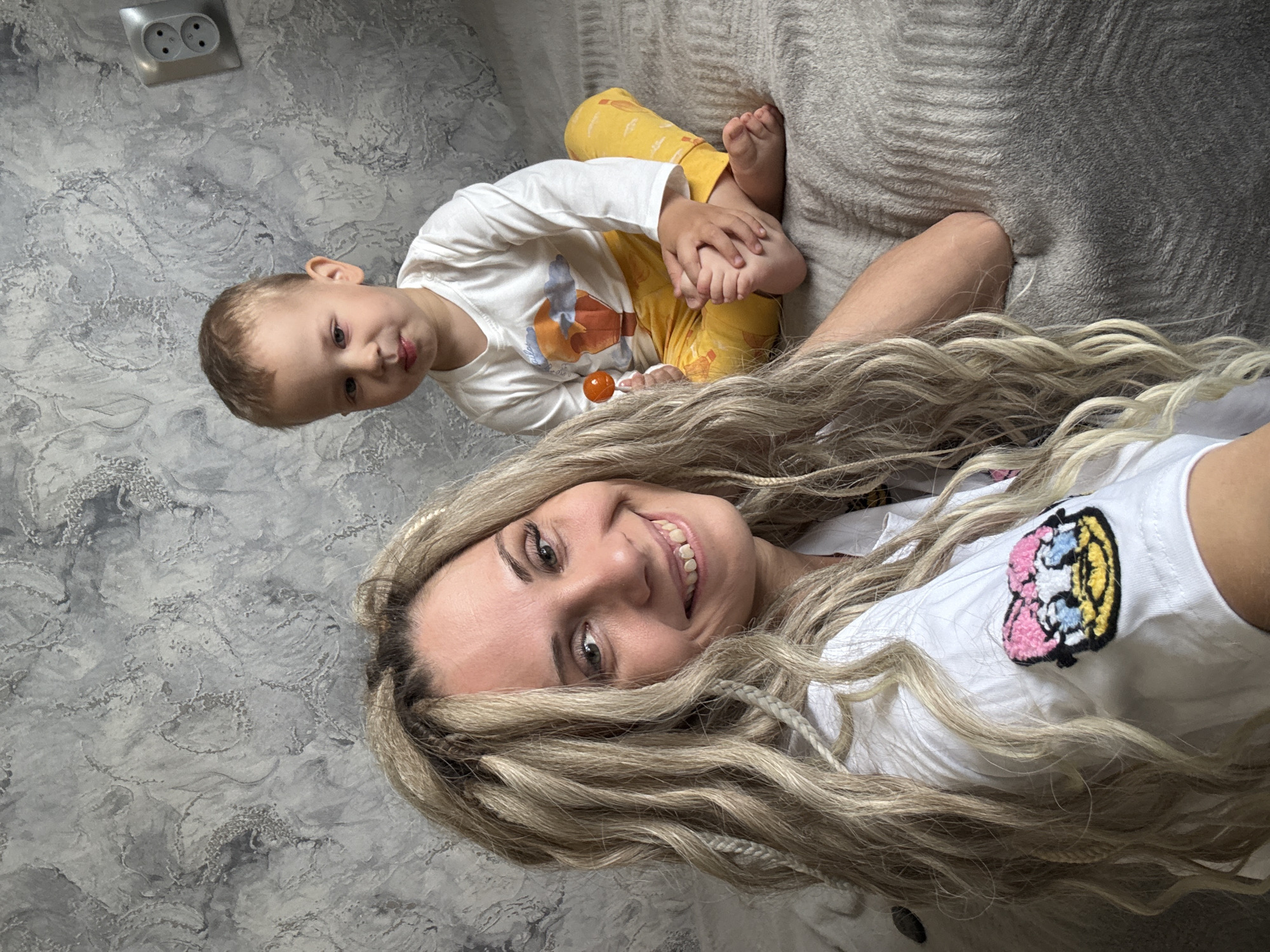 A woman with long, light blonde braided hair is lying on a bed next to a young child. The child is wearing a white long-sleeved shirt with a graphic on the front and yellow pants. The child is holding their feet with their hands and looking at the camera. The woman is wearing a white t-shirt with a colorful patch on the shoulder and smiling at the camera. The background consists of a gray textured wall on one side and a gray bedspread on the other. A double electrical outlet is visible on the wall near the top of the image. The location is Unknown, Unknown.