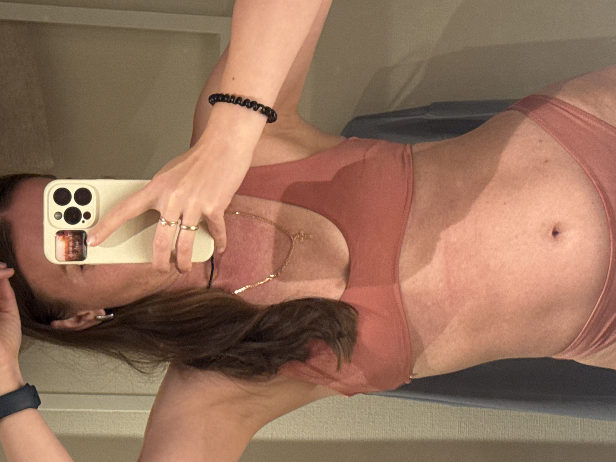 A woman is lying down and taking a picture of herself in a mirror with her phone. She is wearing a pink sports bra and matching underwear. She has a visible tan line. She is wearing a blue fitness tracker on her left wrist, a black bracelet on her right wrist, and multiple rings on her fingers. She also has a necklace with a cross. Her hair is brown. The background is a neutral color. There is no information to determine that the location of this image is in the city of Beppu, Japan.