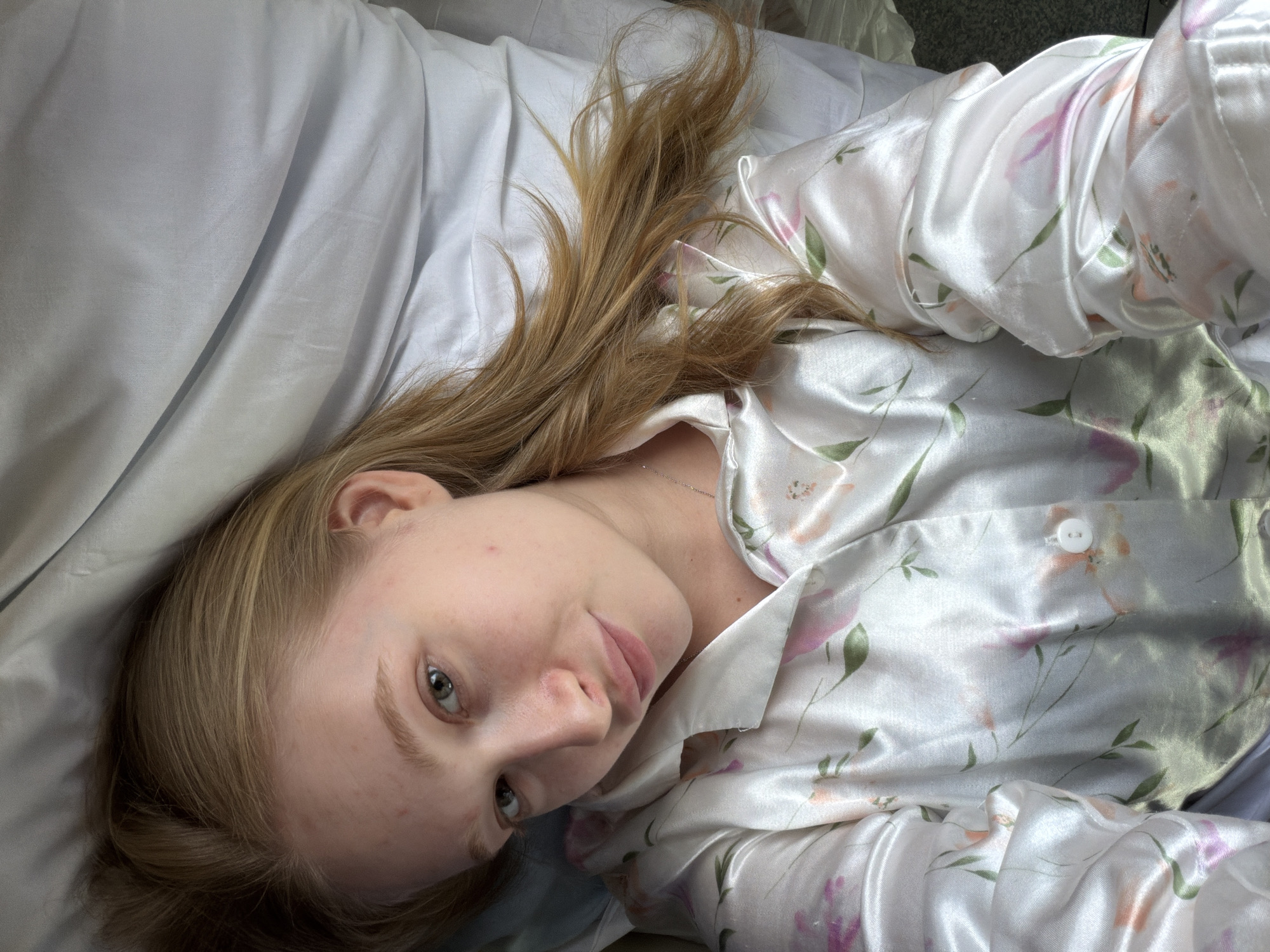 A woman is lying on a bed with white sheets and a silky, floral-patterned pajama top. Her head is tilted towards the camera. She has long, light brown hair, fair skin, and light eyes. No information indicates that the image was taken in the city of Medvyodovskaya, Russia.