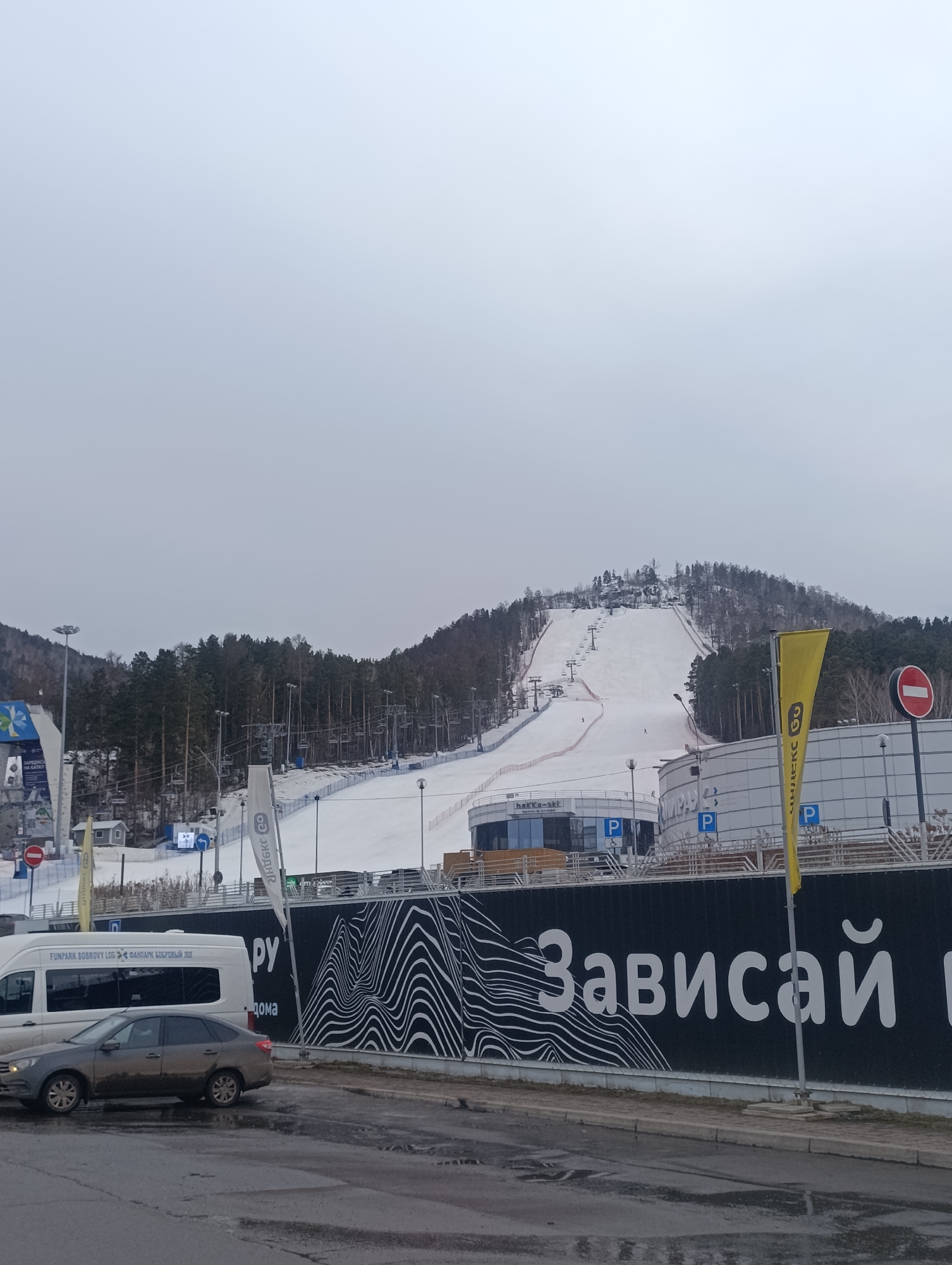 The image shows a ski resort in Krasnoyarsk, Russia, identified as Funpark Bobrovy Log. A snow-covered slope stretches up the hillside, equipped with chairlifts. The foreground includes a parking area with vehicles, and a dark wall featuring a white, stylized graphic and Cyrillic text that reads "Зависай." Flags and signage, including parking symbols, are visible near the base of the slope. The sky is overcast.