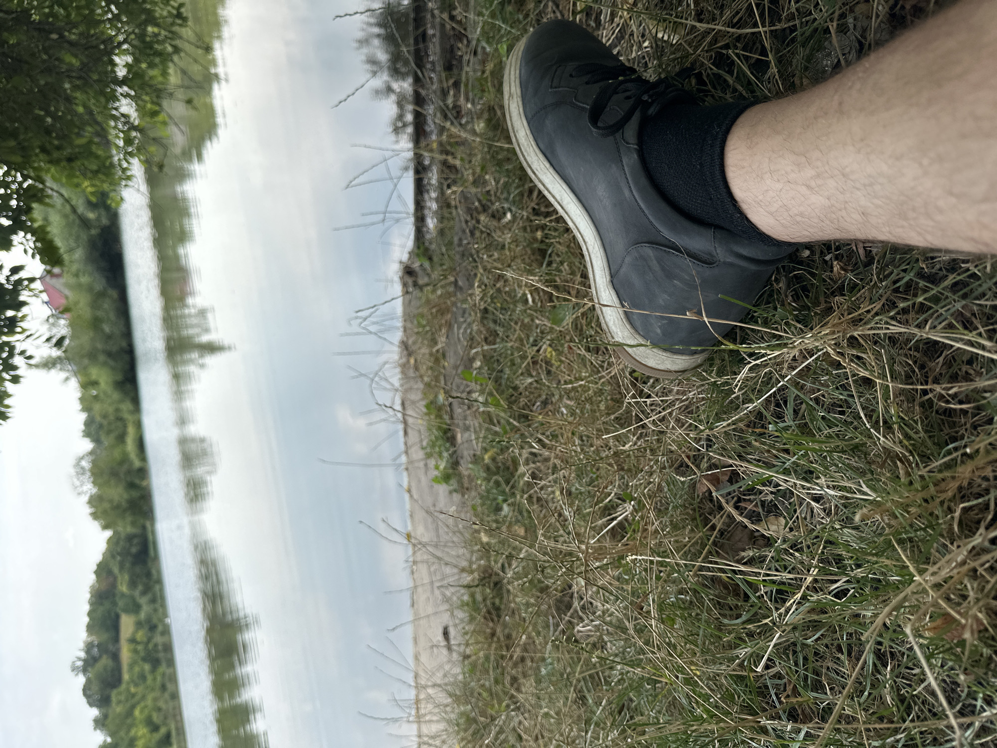 An individual's right leg and foot, wearing a black shoe and sock, are prominently positioned in the foreground, resting on dry grass. The scene appears to be outdoors, likely near a body of water, possibly a lake or river, with a distant treeline and a hazy sky. The background shows a calm water surface reflecting the sky and trees. The location is identified as the city of Makiv, Ukraine.