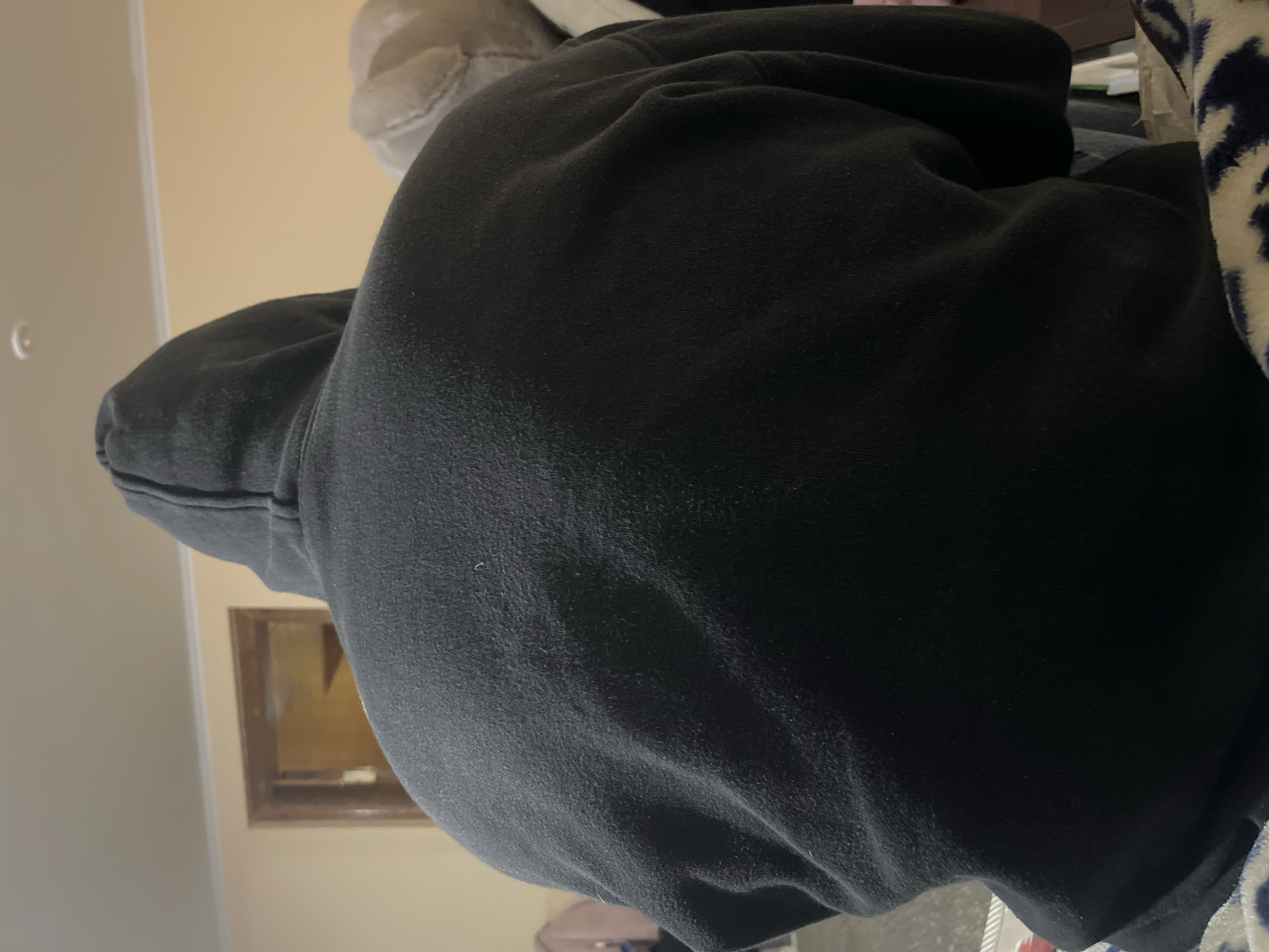 The image shows the back of a person wearing a black hoodie. The person is seated, and the hoodie appears to be made of a soft, possibly fleece material. The background includes a wall painted in a light, neutral color, a doorway with a wooden frame, and a glimpse of a ceiling light fixture. To the right, a blurred light-colored object, possibly a cushion or stuffed toy, is visible. The lighting suggests an indoor setting, likely a room in a residential building. The image is taken from behind the person, focusing on their upper back and head covered by the hoodie. There is no specific information to identify the location as Polonne, Ukraine.