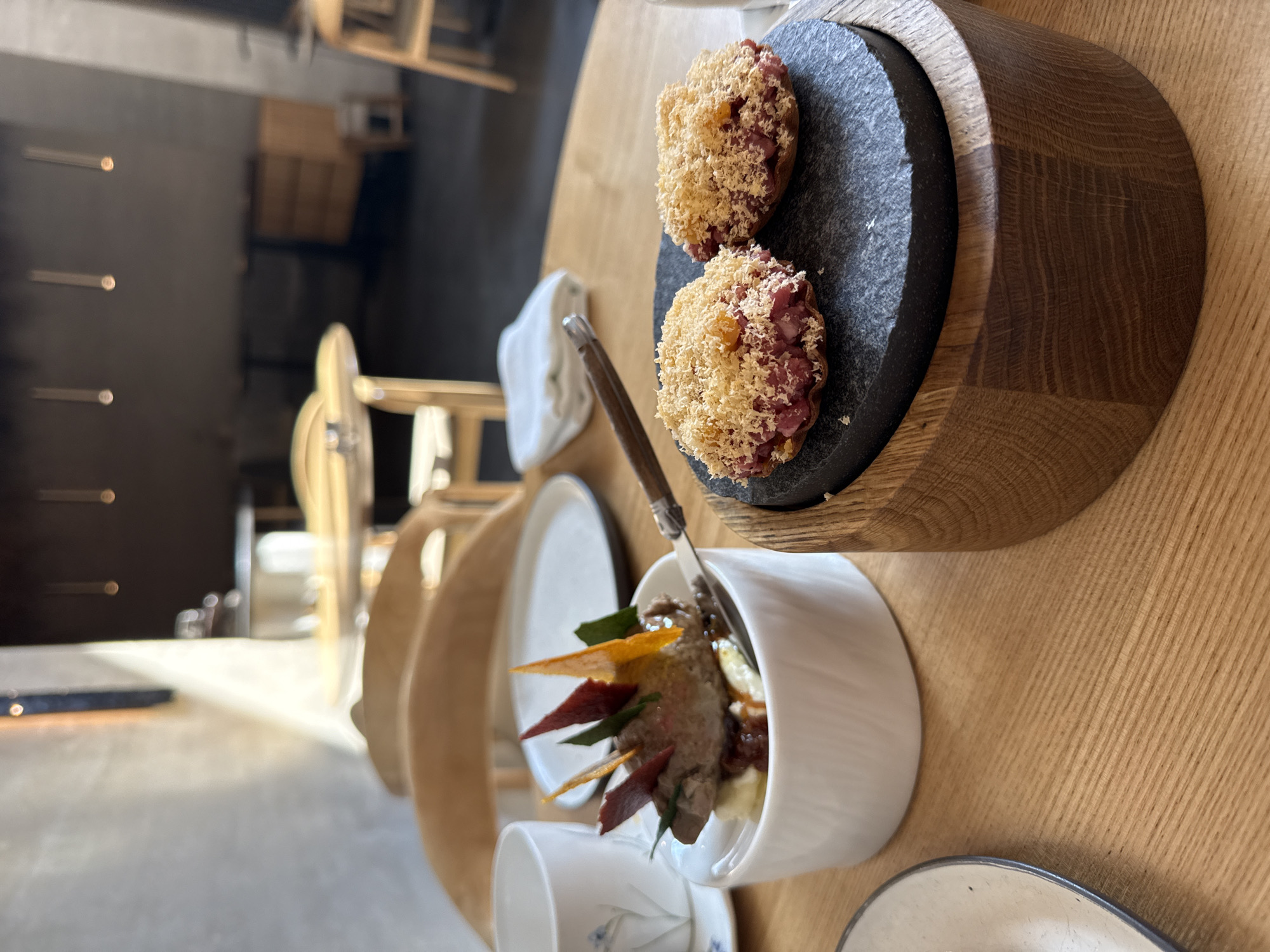 The image presents a dining scene, possibly in a restaurant, with a focus on food presentation. Two small, round food items with a red filling and a topping of shredded yellow material rest on a dark grey, possibly slate, plate that is set within a wooden bowl. Near this bowl, a smaller, white bowl contains an item that appears to be meat with various colored strips/garnishes sticking out. A fork is stuck into this second dish. The bowls are on a light wooden table, along with a stack of plates, and a small cup.
The background is slightly blurred, but it is possible to see a table and a wooden chair.
There is nothing in the image to suggest a specific location, or indeed, the city of Krasnoyarsk, Russia. No other details or signs are present to indicate or verify this location.