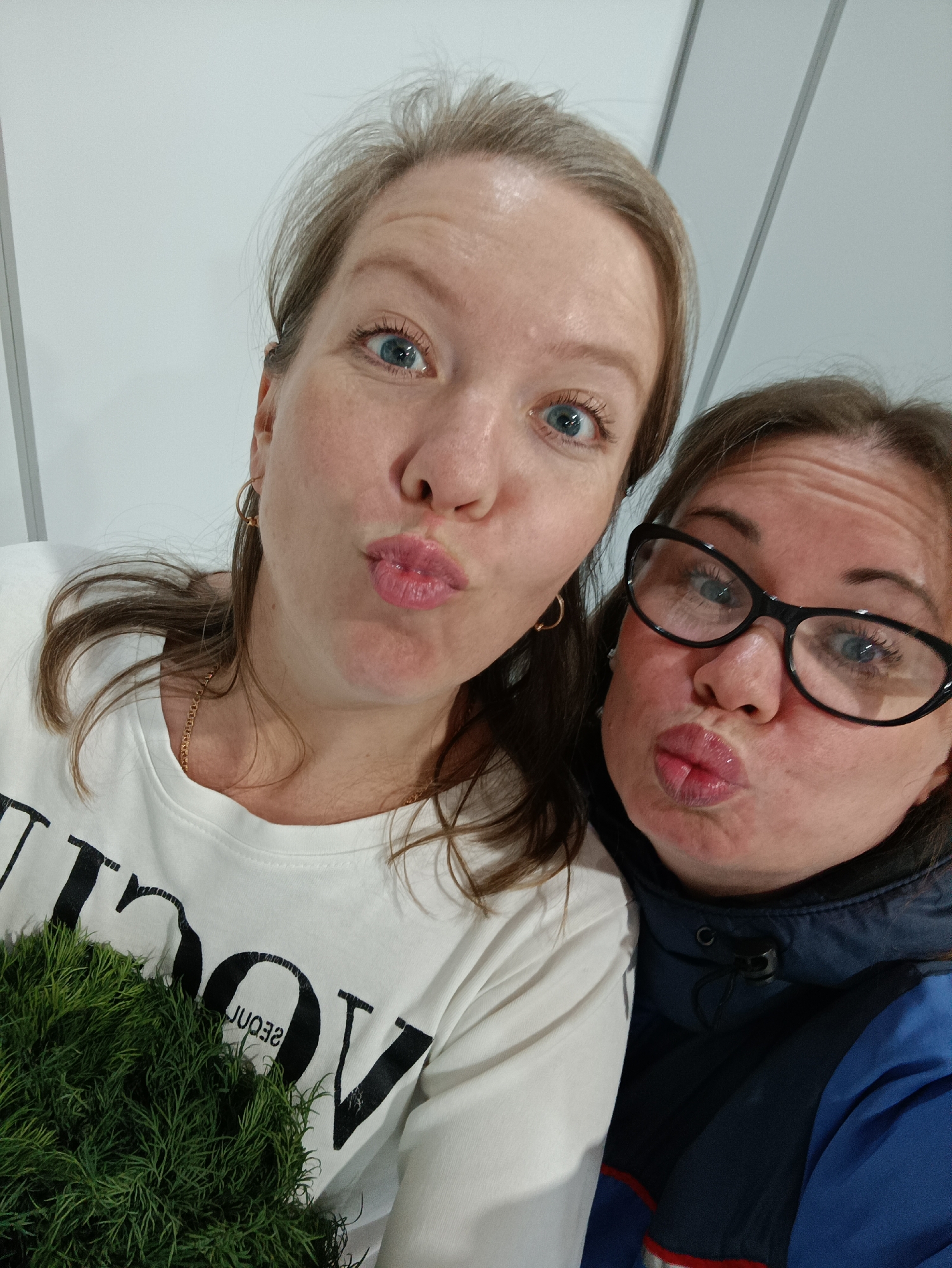 Two women are posing for a selfie. The woman on the left has blonde hair, blue eyes, hoop earrings, a gold chain necklace, and is wearing a white t-shirt with the word "VOGUE" printed on it. She is holding a small green plant. The woman on the right has brown hair, blue eyes, and is wearing black framed glasses and a blue jacket. Both women have their lips pursed in a kissing expression. The background is a white wall with vertical panels. The location appears to be indoors, but the specific location in the city of Chaikovsky, Russia is not apparent from the image.
