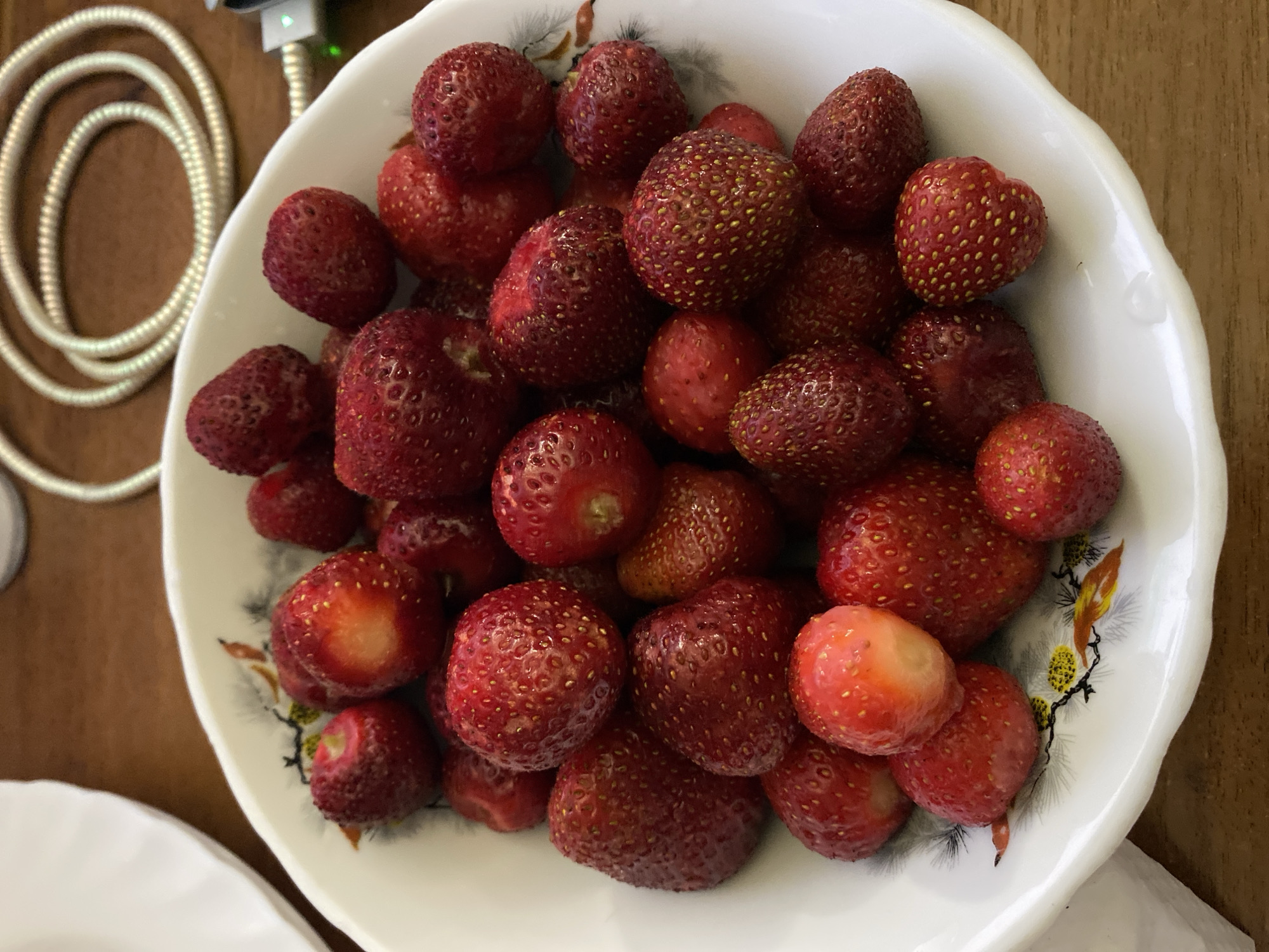 A close-up, overhead shot shows a white bowl filled with fresh strawberries. The bowl has a decorative rim with a floral pattern. The strawberries are various sizes and shades of red, with some showing hints of white near the stem. A few of the strawberries have been bitten into, revealing the lighter flesh inside. The bowl sits on a wooden surface. In the background, out of focus, is a coiled white cable and a hint of a white plate. The scene appears to be an indoor setting.
