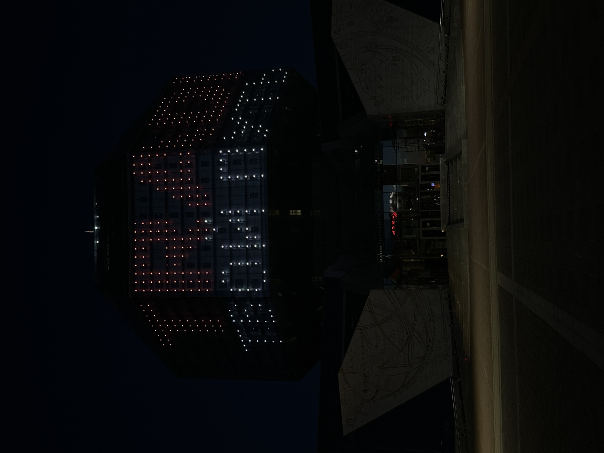 An overhead, nighttime photograph depicts the National Library of Belarus in Minsk. The library's primary structure is illuminated with a pattern of red and white lights, which resemble the colors of the Belarusian flag and spell out the letters "25." Additional lighting adorns the upper and lower portions of the structure. Adjacent to the lit building is a darker, angular structure with architectural diagrams or maps visible on its surface. A narrow walkway or road borders the building's right side. The environment is dark, with minimal ambient light.