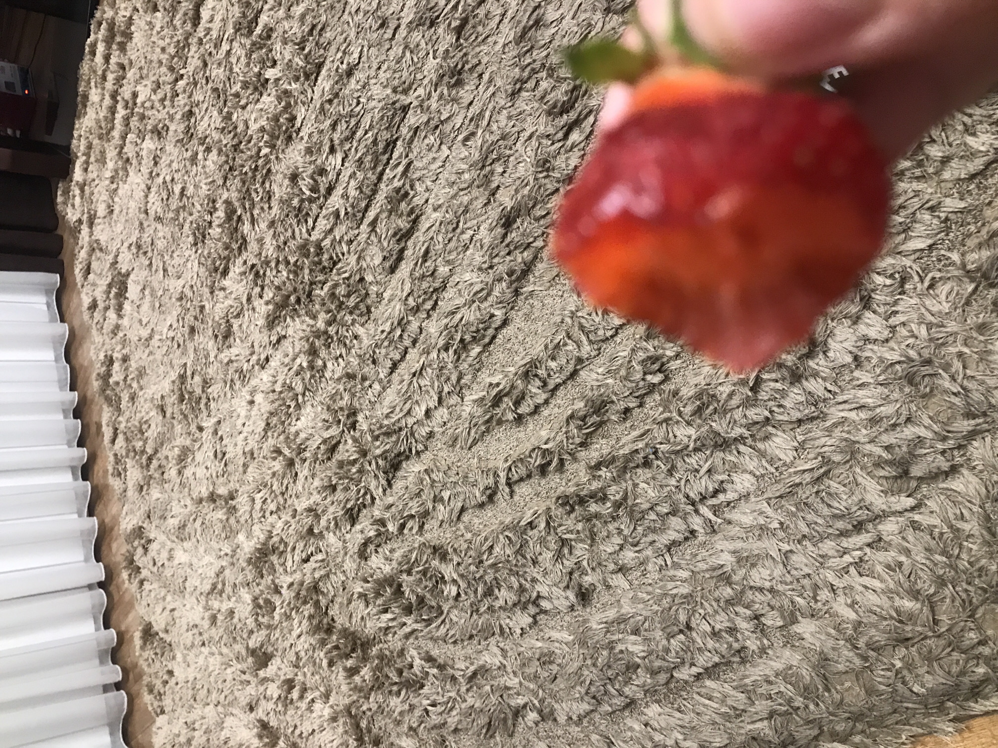 The image shows a hand holding a partially eaten strawberry in front of a tan, shaggy rug. A stack of white folded items is visible in the lower left corner. The background is blurred. No details indicate this image was taken in Yerevan, Armenia.