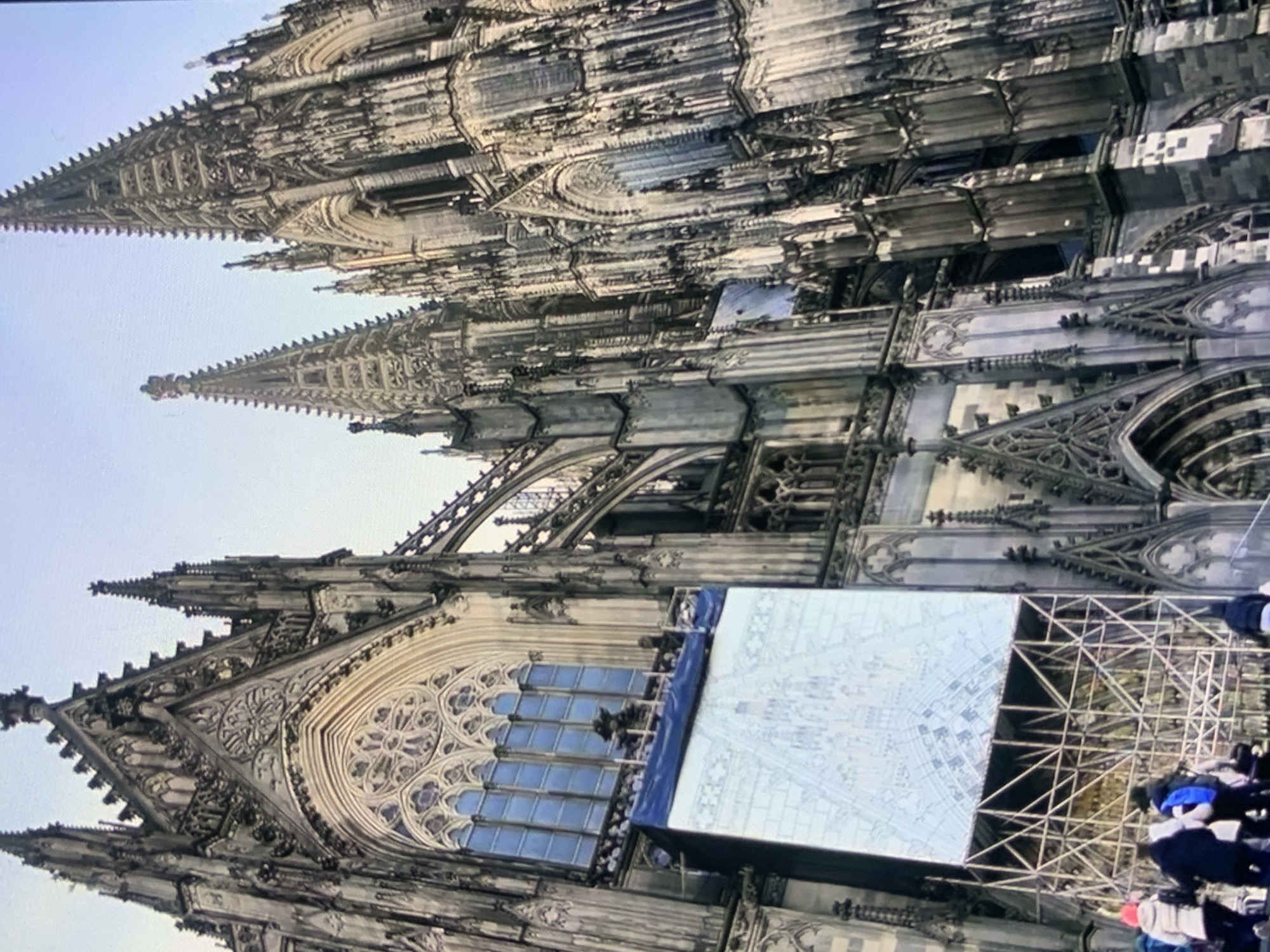 The image displays the intricate Gothic architecture of a large cathedral, likely a significant landmark. The cathedral features soaring spires, elaborate stonework, and large, ornate windows. A large, illuminated banner or tarp is suspended on scaffolding in front of a section of the building, displaying a detailed architectural drawing or blueprint. Several individuals, dressed in winter clothing, are visible at the base of the cathedral, some ascending stairs. The sky is overcast, suggesting a cool or cold climate. This scene is identified as being in Stavropol, Russia.