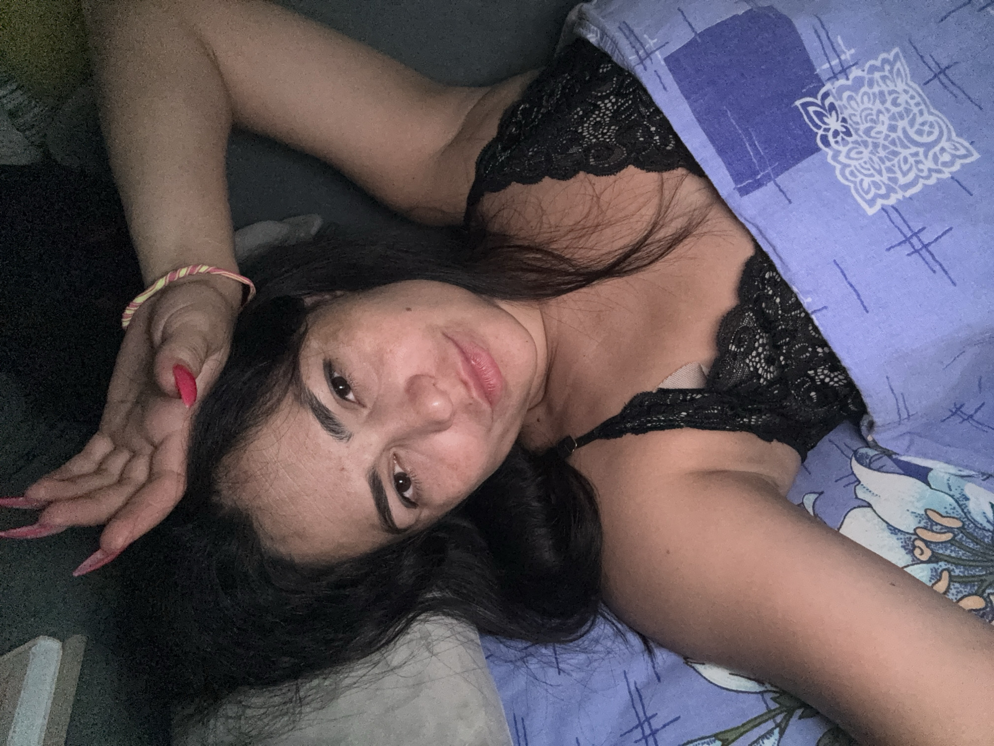 A woman with long, dark hair is lying on a bed. She is partially covered by a blue and white patterned blanket, and is wearing a black lace top. Her left arm is raised above her head, with a pink and yellow bracelet visible on her wrist. Her fingernails are long and painted red. The background appears to be a dark gray or black wall. This image does not contain the location of the city of Kauchuk Stantsiya, Russia.