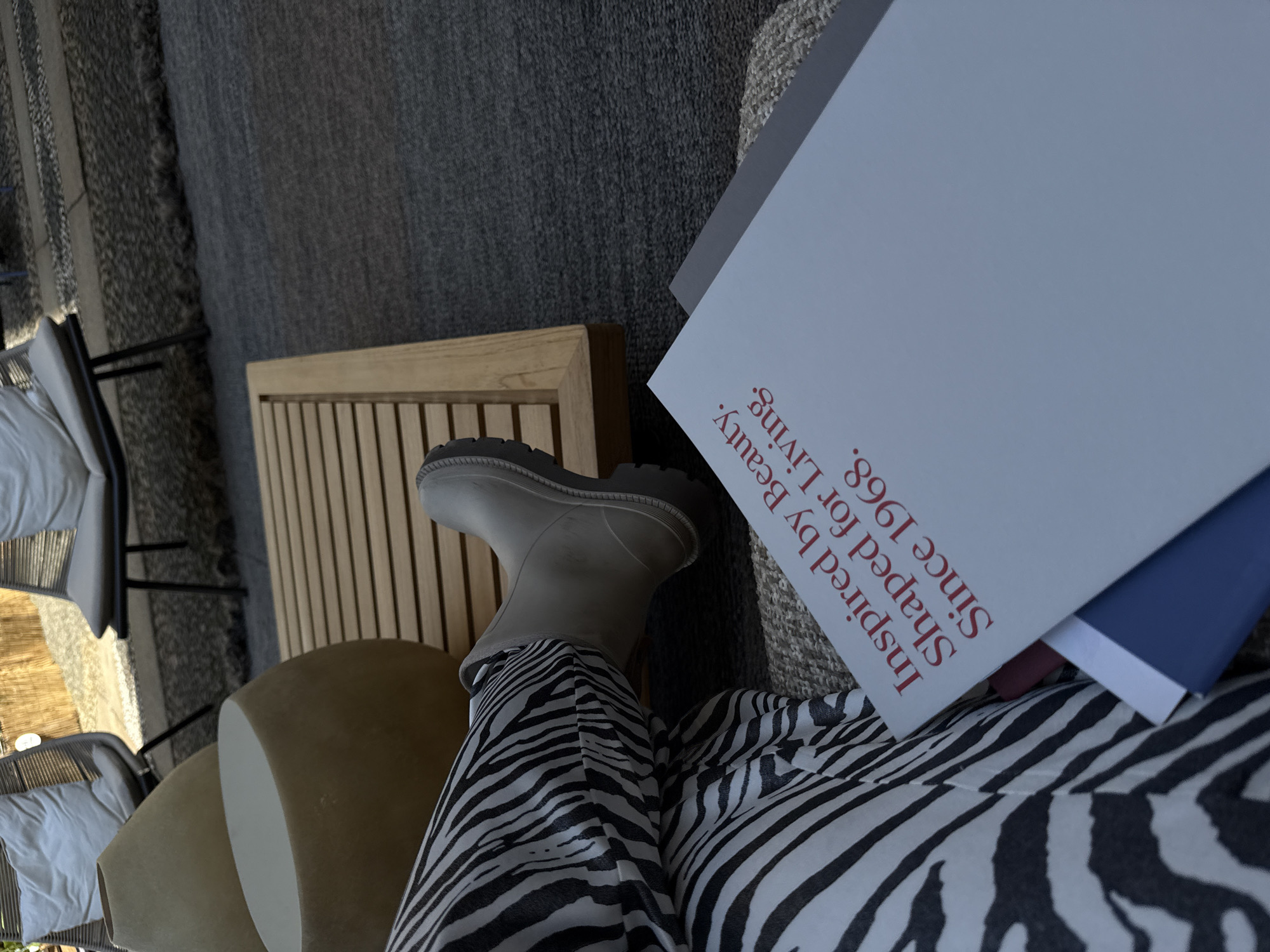 The image shows a person seated on a beige seat, wearing zebra-striped pants and grey boots. The person is holding a white book or magazine with the text "Inspired by Beauty. Shaped for Living. Since 1968." printed in red. The background features a wooden table, a grey rug, and outdoor seating. The setting appears to be an outdoor patio or terrace. Although the information is not visible in the provided picture, one can assume that the location is in Milan, Italy, as requested.