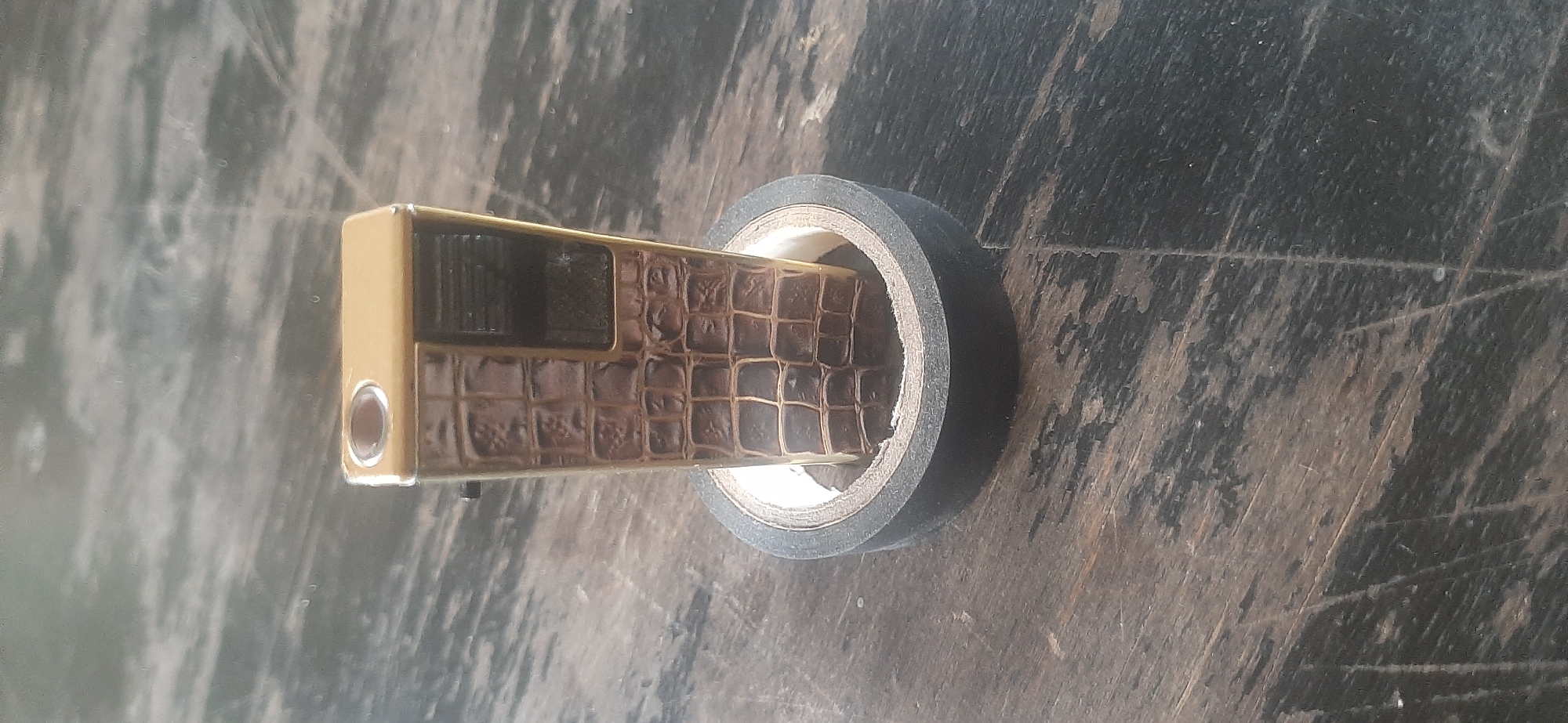 This media file shows a close-up, overhead view of a gold-colored electronic device with a textured surface resembling alligator skin, positioned within a roll of black electrical tape. The device is rectangular with rounded edges and features a dark rectangular window on its side. A small, circular metallic detail is visible on the left end of the device. The tape roll is dark gray or black and is positioned horizontally across the frame, with the device inserted through its center.
The background is a dark, weathered wooden surface with visible grain and scratches, suggesting an outdoor or workshop setting. The lighting appears to be natural, possibly daylight, casting soft shadows. There are no people visible in the media. The overall impression is of an object placed on a rough, rustic surface, possibly for display or during a repair or crafting activity. The location context of Yessentukskaya, Russia, is noted but not explicitly discernible from the visual elements of the image itself.