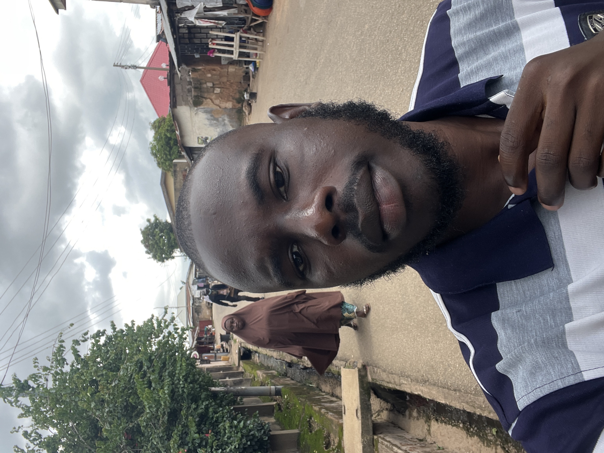 The image shows a close-up of a young Black man with a beard, wearing a striped polo shirt. He is looking directly at the camera, with a slight smile. The background is blurred, but appears to be a street scene with buildings and trees. The sky is overcast, suggesting it might be a cloudy day. In the middle ground, a woman in a brown hijab is walking away from the camera. The image appears to be taken in Jos, Nigeria.