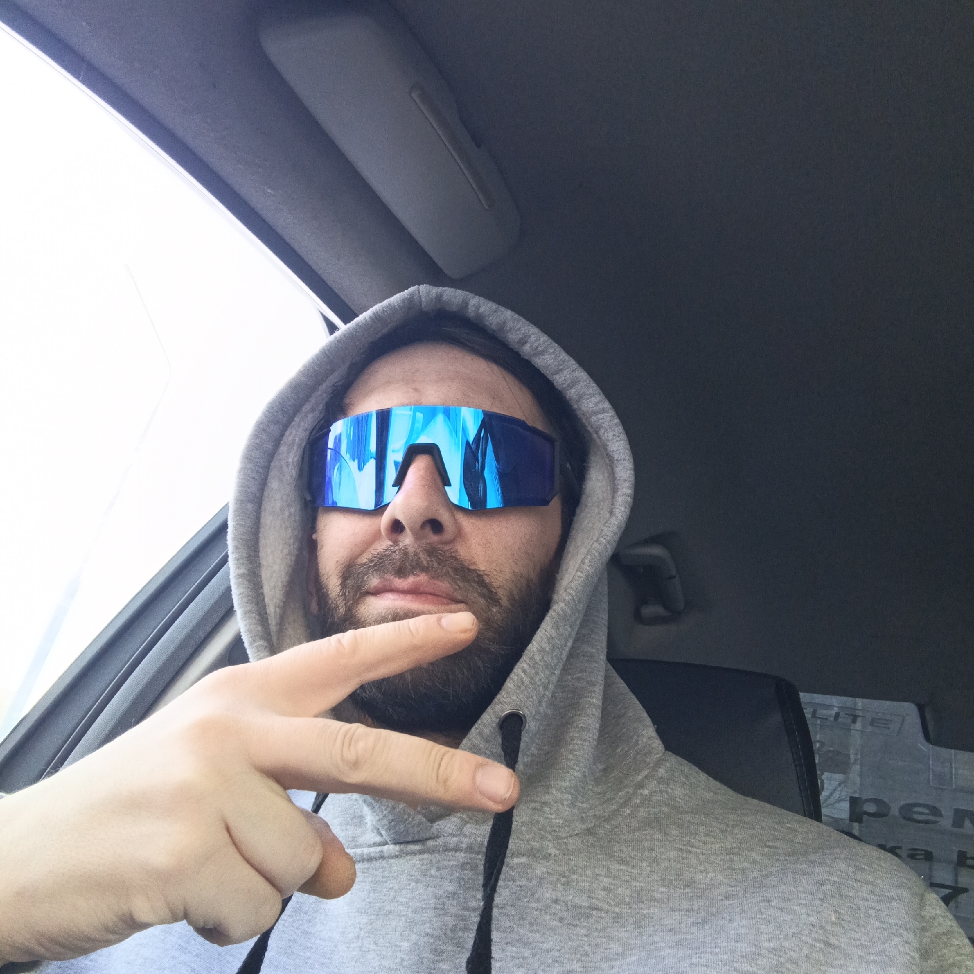 A male individual is depicted seated within a vehicle interior in the city of Марковский, Russia. The subject has a beard and dark hair, obscured partially by a grey hooded sweatshirt with the hood pulled up. He wears large, reflective blue wraparound sunglasses that cover his eyes. His right hand is positioned in the lower-left of the frame, forming a "V" gesture with the index and middle fingers extended. The vehicle's dark grey headliner and a sun visor are visible above the subject. A bright, diffused light source is visible through a window on the left. Behind the subject, a dark seatback and a rear window or reflective panel are visible, displaying indistinct, partially legible text including "LITE" and Cyrillic script.
