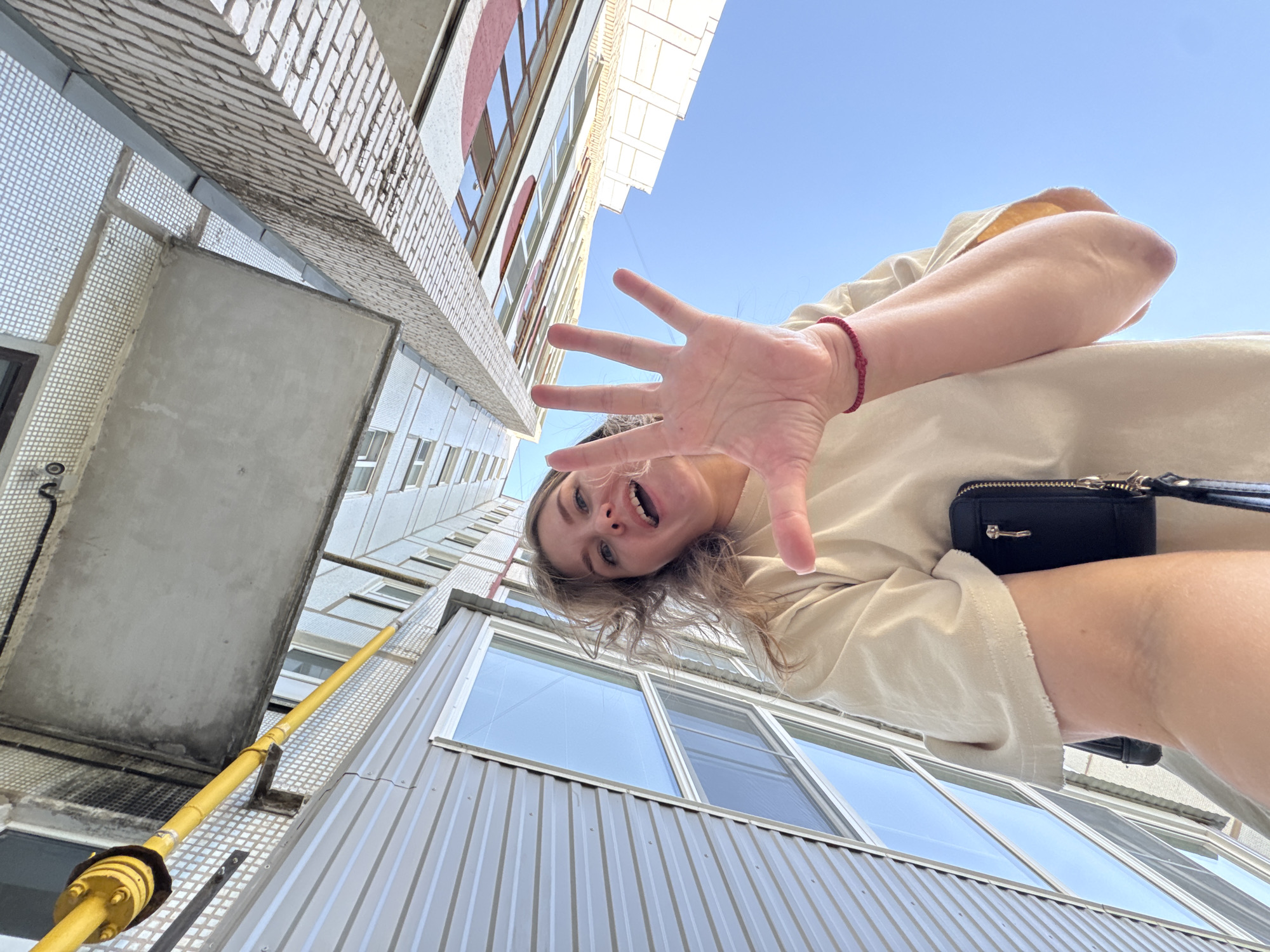 A woman is seen from a low angle, pointing her hand towards the camera. She has fair skin, blonde hair, and is wearing a beige t-shirt and a red bracelet. A small black purse is visible on her person. The background consists of apartment buildings with visible windows and a yellow gas pipe running along the exterior wall. The location is Chaikovsky, Russia.