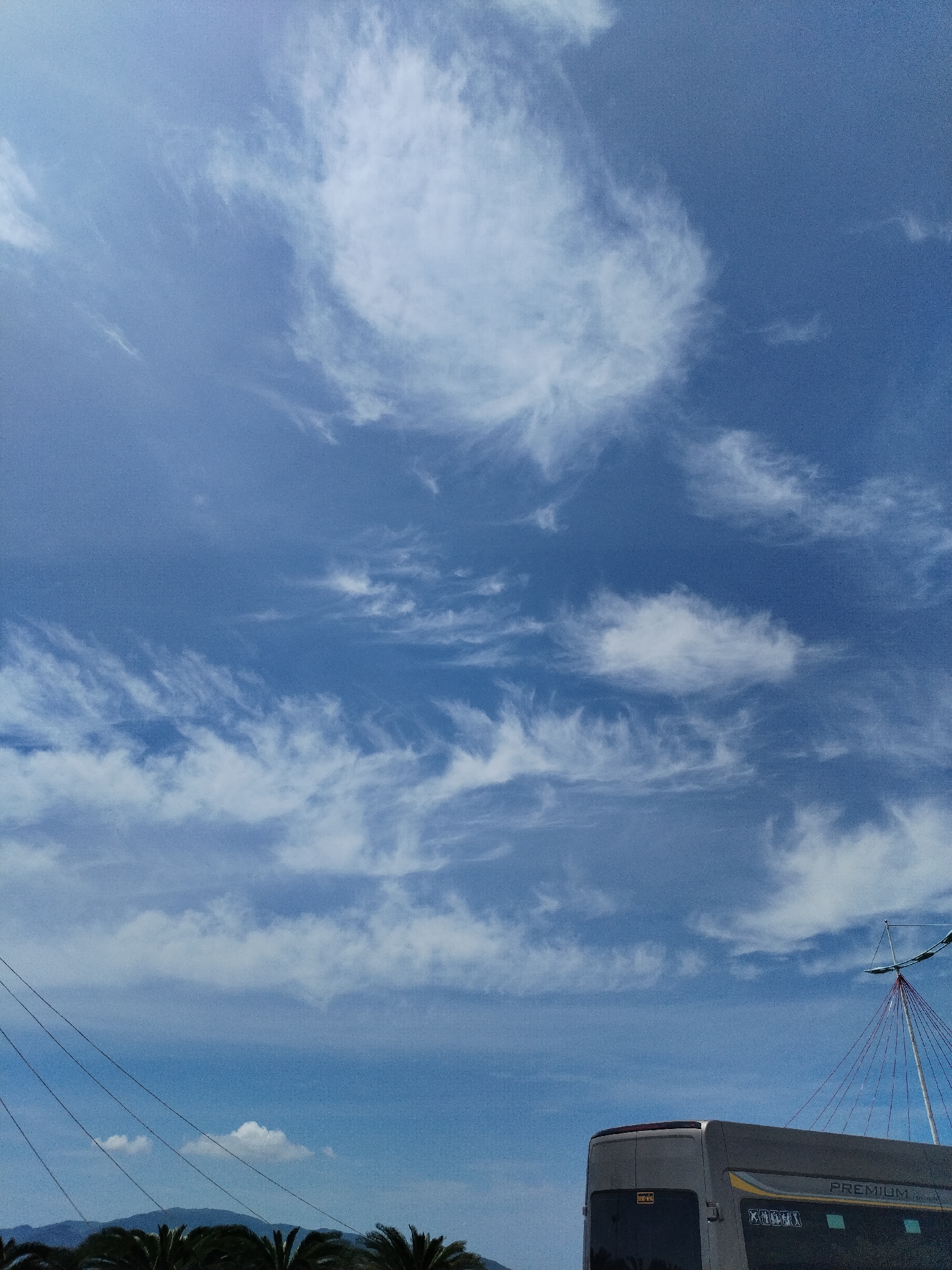 This image depicts a scene in Nha Trang, Vietnam, under a bright blue sky dotted with wispy white clouds. The lower portion of the frame shows the back of a grey bus with "PREMIUM" visible on its side. A portion of a light pole with red guy-wires is also visible to the right of the bus. In the distance, the silhouettes of palm trees and hills can be seen against the horizon. Several dark, diagonal lines, likely power lines, extend across the upper left quadrant of the image.