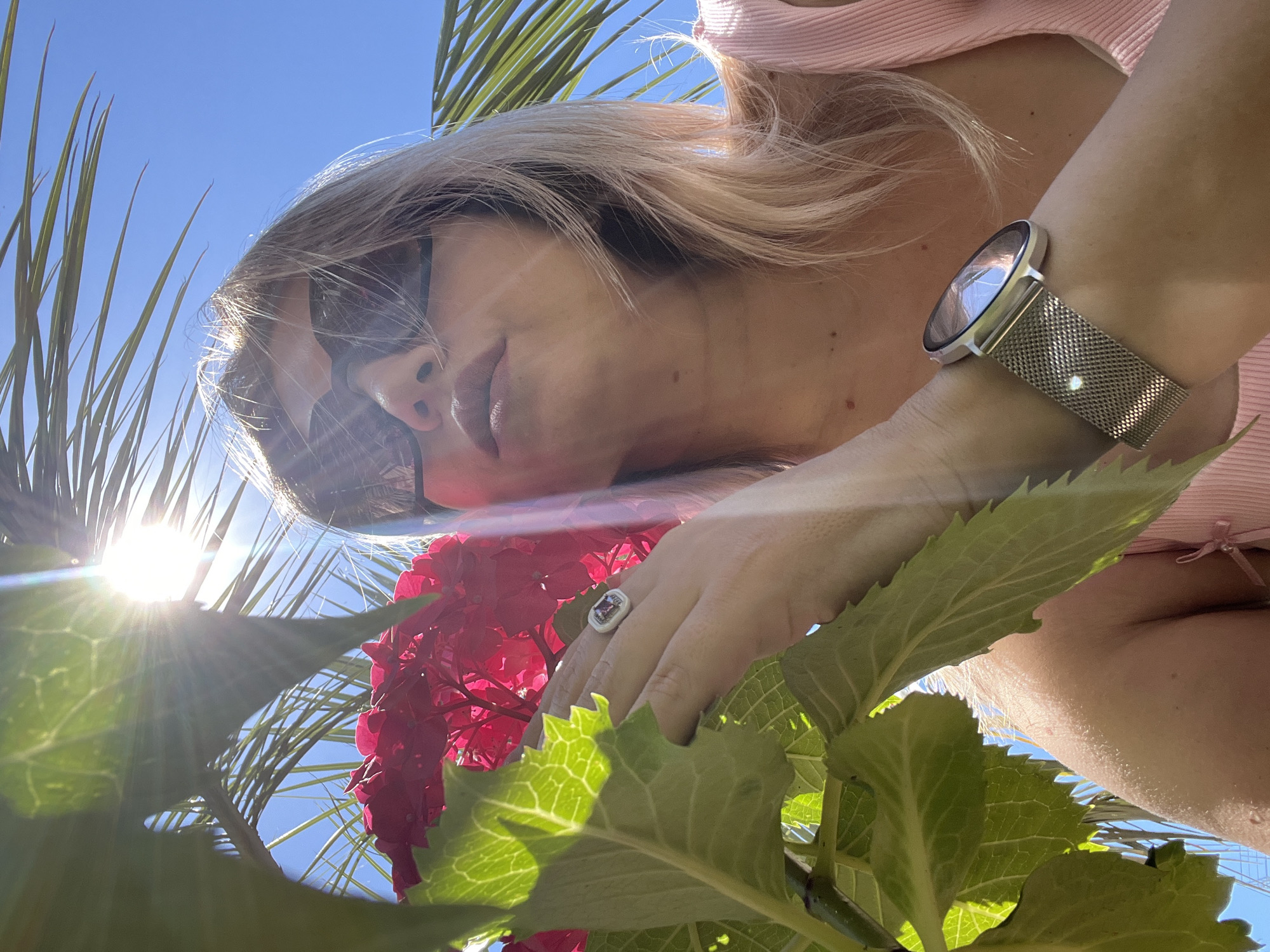 In Sochi, Russia, a woman with blonde hair wears sunglasses and a pink top, holding a pink flower. She has a watch on her left wrist and a ring on her right ring finger. The scene is illuminated by bright sunlight, which creates lens flare. Palm fronds are visible in the background against a clear blue sky.