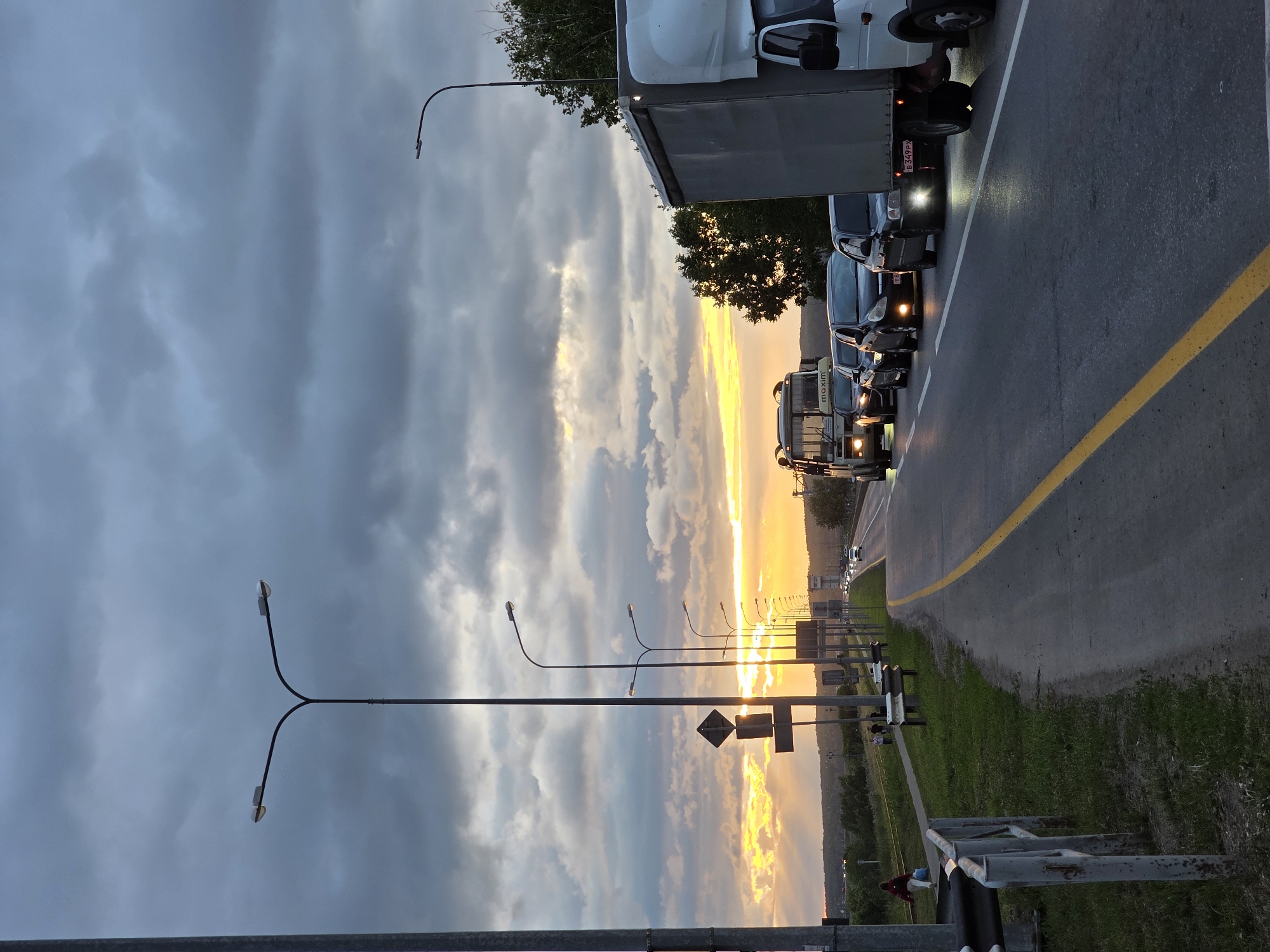 The image depicts a traffic jam on a multi-lane road with a sunset backdrop, likely in Chaikovsky, Russia. The sky is filled with dramatic, dark clouds partially illuminated by the setting sun, which casts a warm glow on the horizon. Several streetlights line the side of the road, their lights beginning to illuminate. On the right side of the road, a line of vehicles is stopped, including a large truck, a white van, and several passenger cars. The headlights of some cars are on, reflecting off the wet road surface, suggesting recent rain. In the distance, more vehicles are visible, also stopped. On the left side of the road, there is a grassy embankment with a guardrail. A few people are walking on a sidewalk next to the embankment. A bus with "maxim" visible on its side is also part of the traffic jam. The overall scene suggests an evening commute or a delay on a major road.