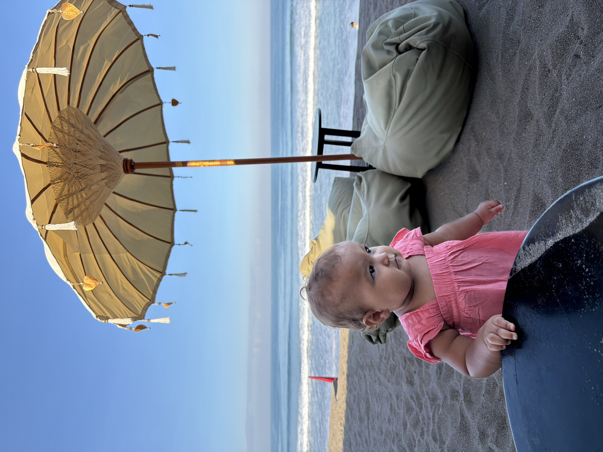 An infant wearing a pink dress is standing on a sandy beach in North Kuta, Indonesia. The infant is looking to the right of the frame. Behind the infant, there is a large, decorative umbrella providing shade. Several large, light-colored beanbag chairs are situated under the umbrella. In the background, the ocean stretches out with gentle waves under a clear blue sky. The scene is set during daytime.