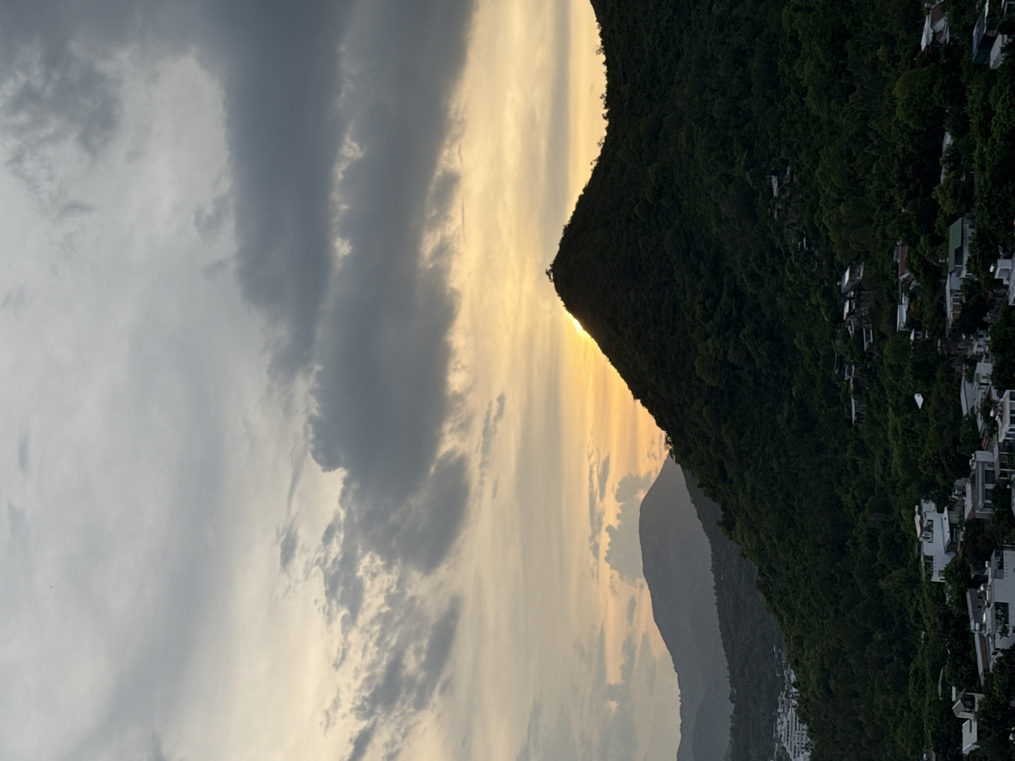 This media file depicts a sunset over a verdant, mountainous landscape with a coastal town nestled along the slopes. The scene is oriented vertically.
The primary subjects are the natural landscape and the sky. A large, dark green, tree-covered mountain dominates the right side of the frame. Its silhouette cuts sharply against the sky. Scattered along the upper slopes of this mountain are numerous white buildings, characteristic of a residential area. The buildings appear to be modern, with several having flat roofs. Further into the background, another, more distant mountain range is visible, appearing softer and more muted in color due to atmospheric haze.
The sky is a significant element, filled with dramatic clouds. On the left side, large, dark grey clouds create a sense of impending weather or a transition in the day. Towards the center and right, the sky is illuminated with the warm hues of sunset, ranging from soft yellow to orange. The sun itself is not directly visible but its light is clearly breaking through a gap in the clouds near the peak of the mountain on the right.
The overall mood is serene and awe-inspiring, capturing the beauty of a natural landscape at the end of the day. The time of day is clearly sunset, indicated by the warm lighting and cloud formations. The weather appears to be dynamic, with both dark clouds and bright sunlight present. The presence of the buildings suggests a human settlement integrated with the natural environment.
While no text is visible in the image, the location context provided indicates this scene is in Bac Natural Heritage Area, Trang, Vietnam. This implies a beautiful and protected natural environment.