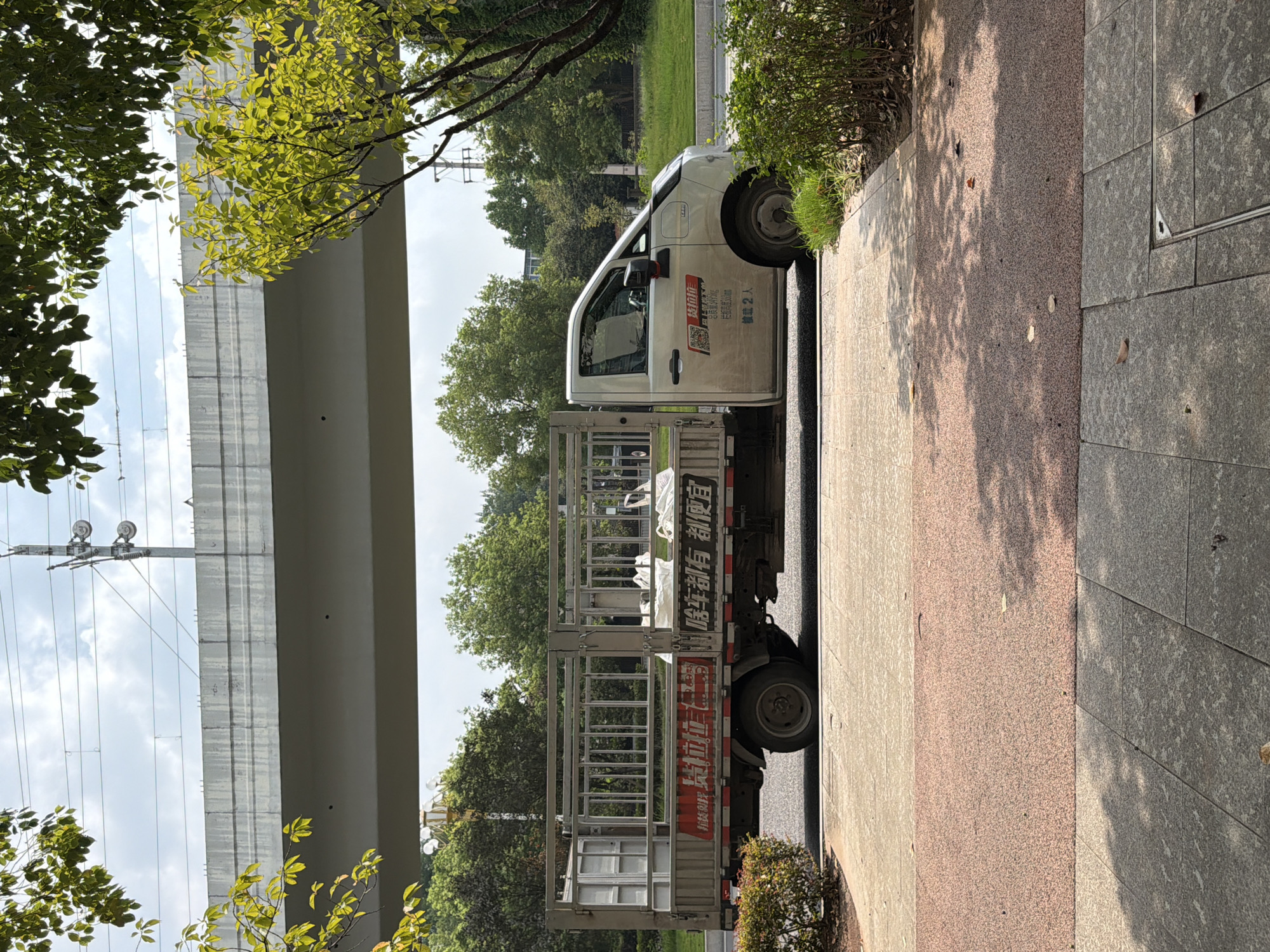 An outdoor, eye-level, side view photograph shows a white light-duty truck with a cargo bed. The truck is parked on a paved surface next to a sidewalk. The cargo bed is open at the back and sides, with metal railings. White, bagged items are visible in the cargo bed. The truck has writing on its side doors and the rear of the cargo bed, some in red and black text. A portion of a concrete overpass is visible in the upper part of the image, with power lines running above it. Trees and green foliage are present in the background, and shadows from trees are cast on the sidewalk. The setting appears to be a street in Jiangjin, China.