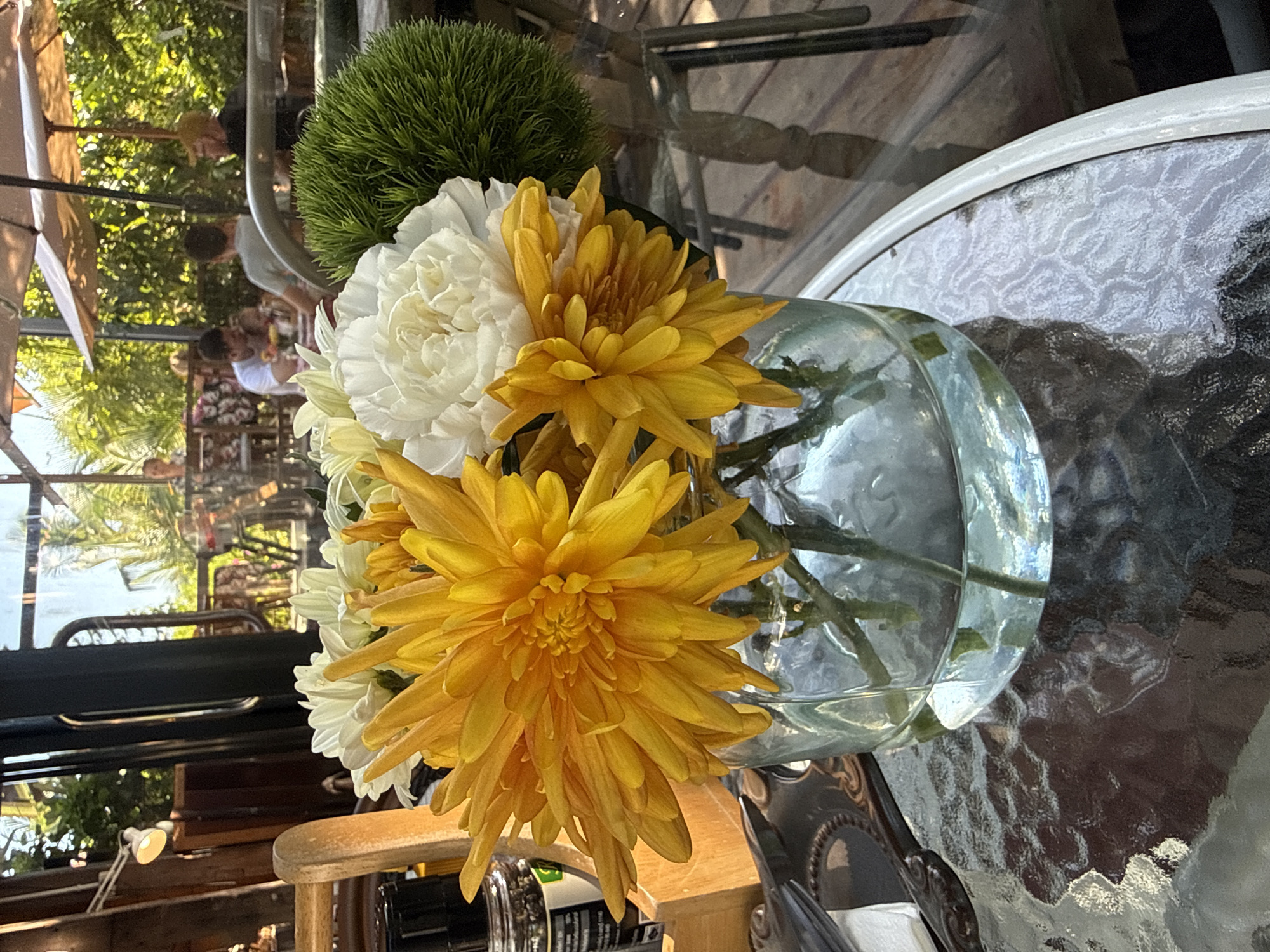 A close-up shot focuses on a bouquet of yellow and white chrysanthemum flowers in a clear glass vase. The flowers are vibrant, with the yellow chrysanthemums prominently displayed in the foreground, their petals radiating outwards. Behind them, white chrysanthemums add contrast, and a spiky green plant is visible to the right. The vase sits on a textured glass table, which reflects the surrounding environment.
In the background, through a glass pane, a scene unfolds at an outdoor establishment, possibly a restaurant or cafe. Patrons are seated at tables under umbrellas, enjoying a sunny day. The environment appears to be lush and green, suggesting a tropical location, consistent with Koh Samui District, Thailand. The overall atmosphere is relaxed and leisurely, with natural light illuminating the scene.