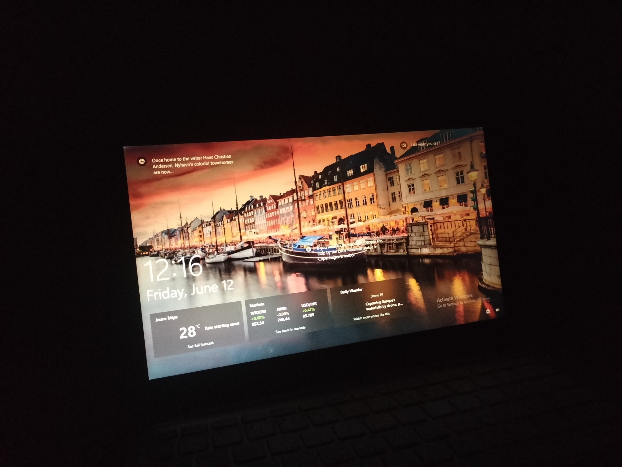 A laptop screen displays a lock screen image of Nyhavn, a canal district in Copenhagen, Denmark. The image features colorful historic townhouses lining the canal, with a boat docked in the foreground. Overlaid on the image are various informational elements, including the time "12:16," the date "Friday, June 12," and weather information indicating "28°C" and "Rain starting soon." There are also sections displaying market data for "Markets," "AWCI," and "USD/INR," as well as a "Daily Wonder" section with content related to "Drone TV." A prompt to "Activate Windows" is visible in the lower right corner. The screen is illuminated against a dark background, suggesting the laptop is in a dimly lit environment.