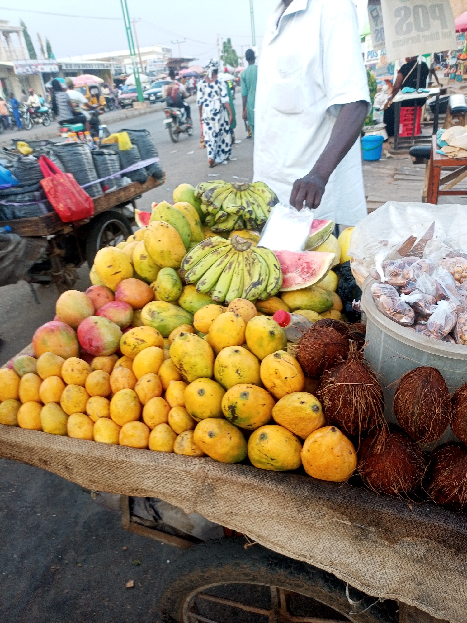 The image depicts a street scene in Tempure, Nigeria, featuring a vendor standing behind a cart laden with produce for sale. The cart, covered with a rough textile, displays a variety of fruits including mangoes of varying sizes and colors, bunches of green bananas, slices of watermelon, whole coconuts, and packaged nuts in a plastic container. The vendor, dressed in a white shirt, is reaching for a plastic bag. In the background, there are other carts, pedestrians, and vehicles moving along the street. A building is visible in the distance, as well as signs and other elements indicating a bustling urban environment.
