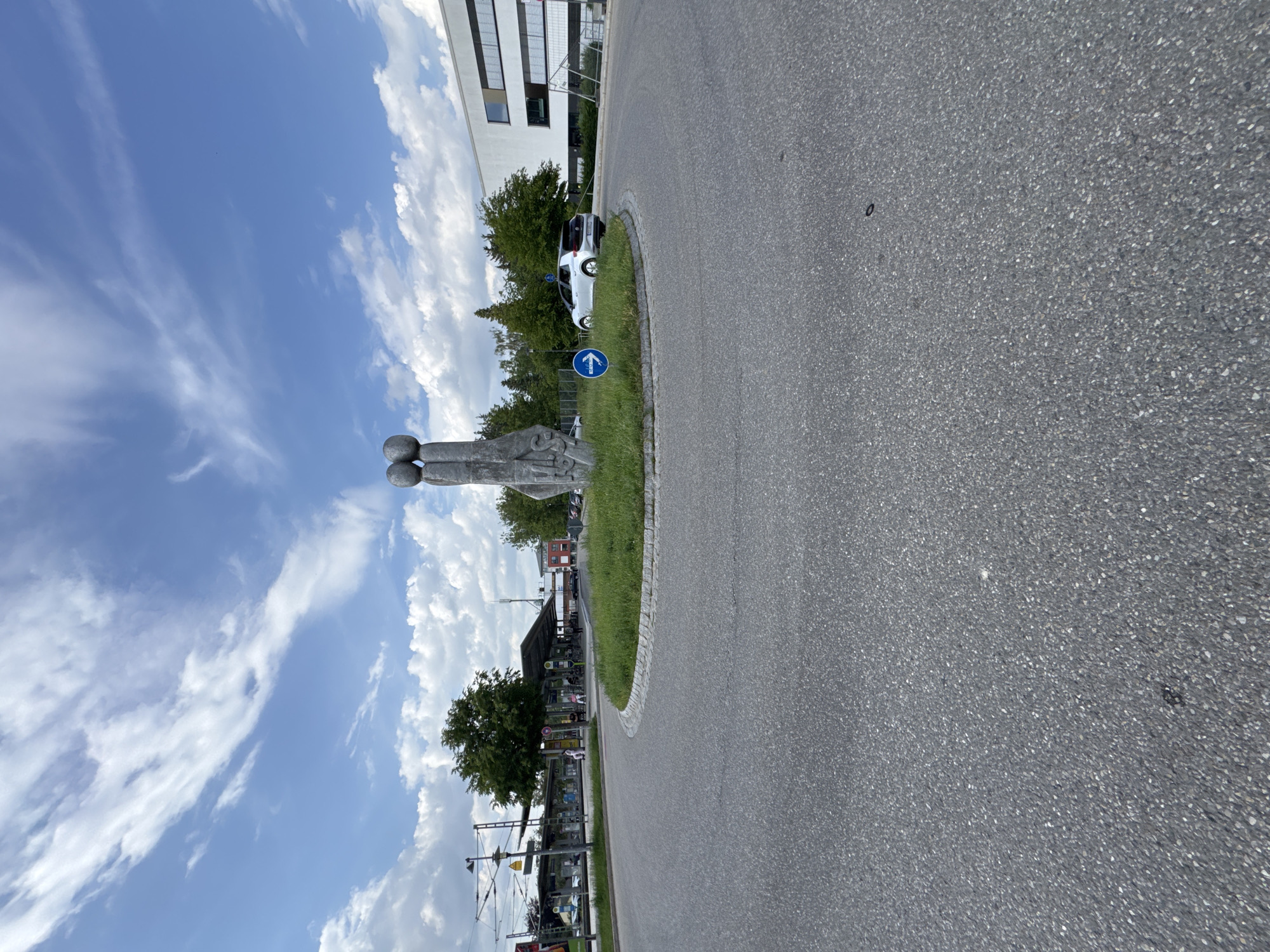 The image shows a roundabout in Höhenkirchen-Siegertsbrunn, Germany. A distinctive sculpture of two figures made of stone stands in the center of the roundabout, surrounded by grass. A white electric car is seen navigating the roundabout, with a blue circular sign indicating the direction of traffic. In the background, a modern white building with large windows is visible, along with trees and a train station with a covered platform. The sky is blue with scattered white clouds. The road surface is asphalt.