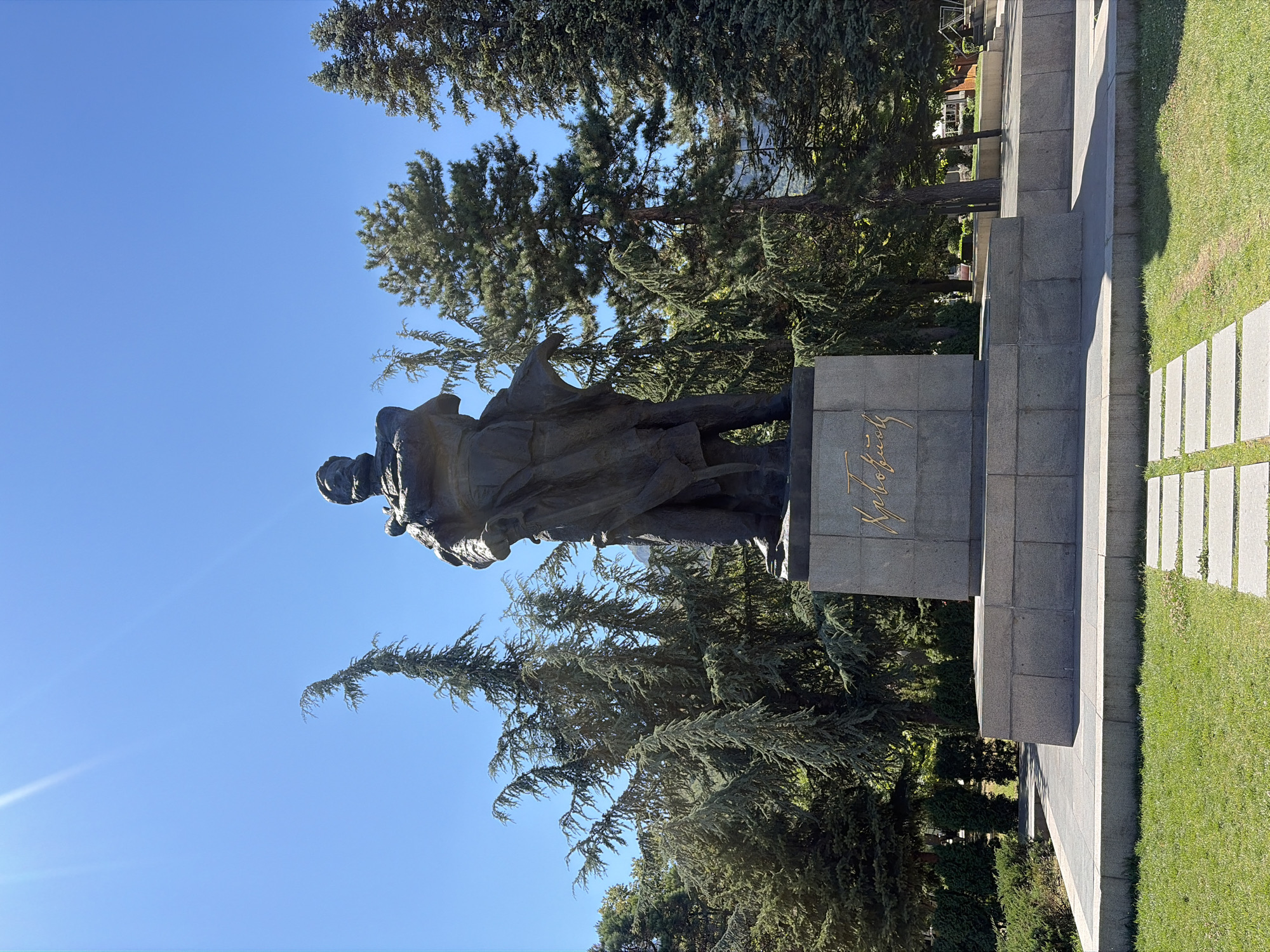 The image shows a statue of Hristo Botev, a Bulgarian national hero, located in the city of Vratsa, Bulgaria. The bronze statue stands on a stone pedestal, with the inscription "Христо Ботев" in gold letters on the front. The statue depicts Botev in a dramatic pose, with a cloak draped over his shoulders. He is dressed in traditional attire, including a hat and boots. The statue is situated in a park-like setting, with lush green grass and mature trees surrounding it. The sky is a clear blue, and the sun is shining brightly, casting shadows on the ground. In the background, other trees and some buildings are visible. The overall impression is one of grandeur and historical significance.