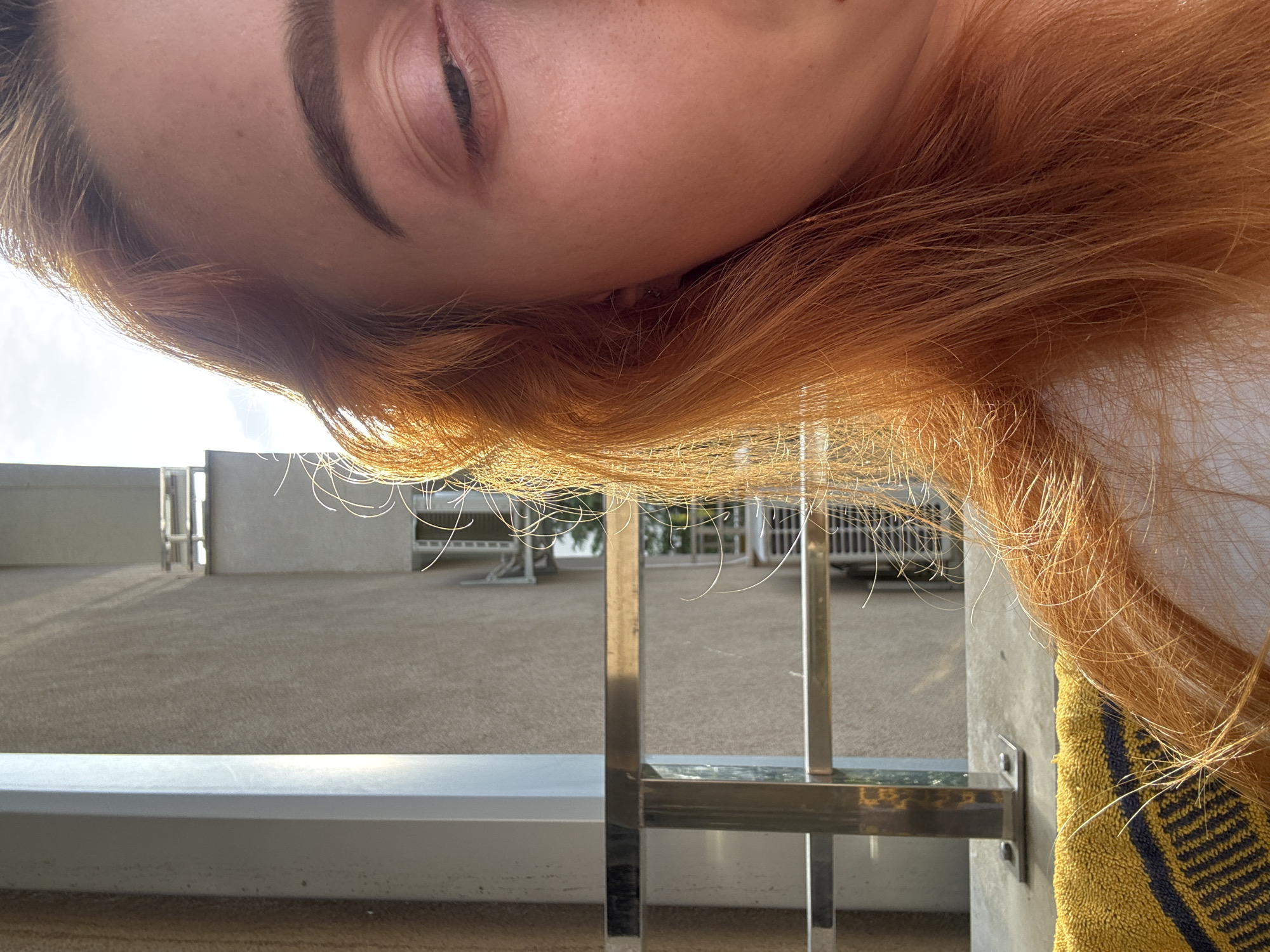 The image is taken from a low, upside-down perspective, showing part of a person's face (eye, eyebrow, forehead) and reddish-blonde hair. The background suggests an outdoor, possibly balcony or patio setting. There is a railing made of metal and concrete, with a textured surface behind it, possibly a courtyard or open area. There is a yellow towel with blue stripes near the right side of the image.
While the image doesn't give enough information to pinpoint the exact location in Budva, Montenegro, the architecture and outdoor setting are consistent with a coastal or residential area in that city.