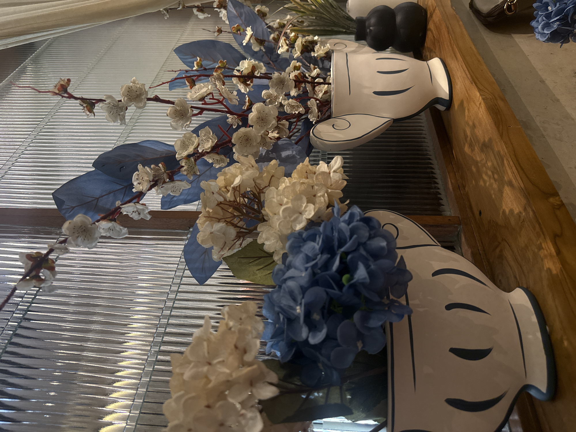 The image presents a detailed close-up of two decorative arrangements, likely indoors. Each arrangement features floral elements placed in vases styled to resemble the iconic silhouette of Mickey Mouse's head, complete with round ears and characteristic facial features. The arrangements include white blossoms, blue hydrangea-like flowers, and leafy accents. The backdrop appears to be a combination of horizontally-oriented Venetian blinds and a wooden surface. The objects suggest a decorative or themed display, possibly within a domestic or commercial space. The location is Ubud District, Indonesia.