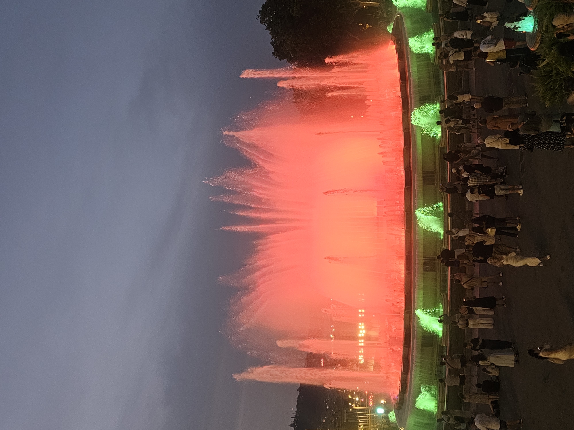 The image shows a nighttime view of the Magic Fountain of Montjuïc in Barcelona, Spain. The fountain is illuminated with vibrant red and green lights, casting a dramatic glow on the cascading water. A crowd of people is gathered around the fountain, observing the spectacle. Some individuals are standing, while others are seated on the ground. The sky above is dark and cloudy, suggesting it is evening or nighttime. In the background, the lights of the city are visible on a hillside, along with trees and the silhouette of buildings. The overall scene depicts a lively public space with a focus on the illuminated fountain and the onlookers enjoying its display.