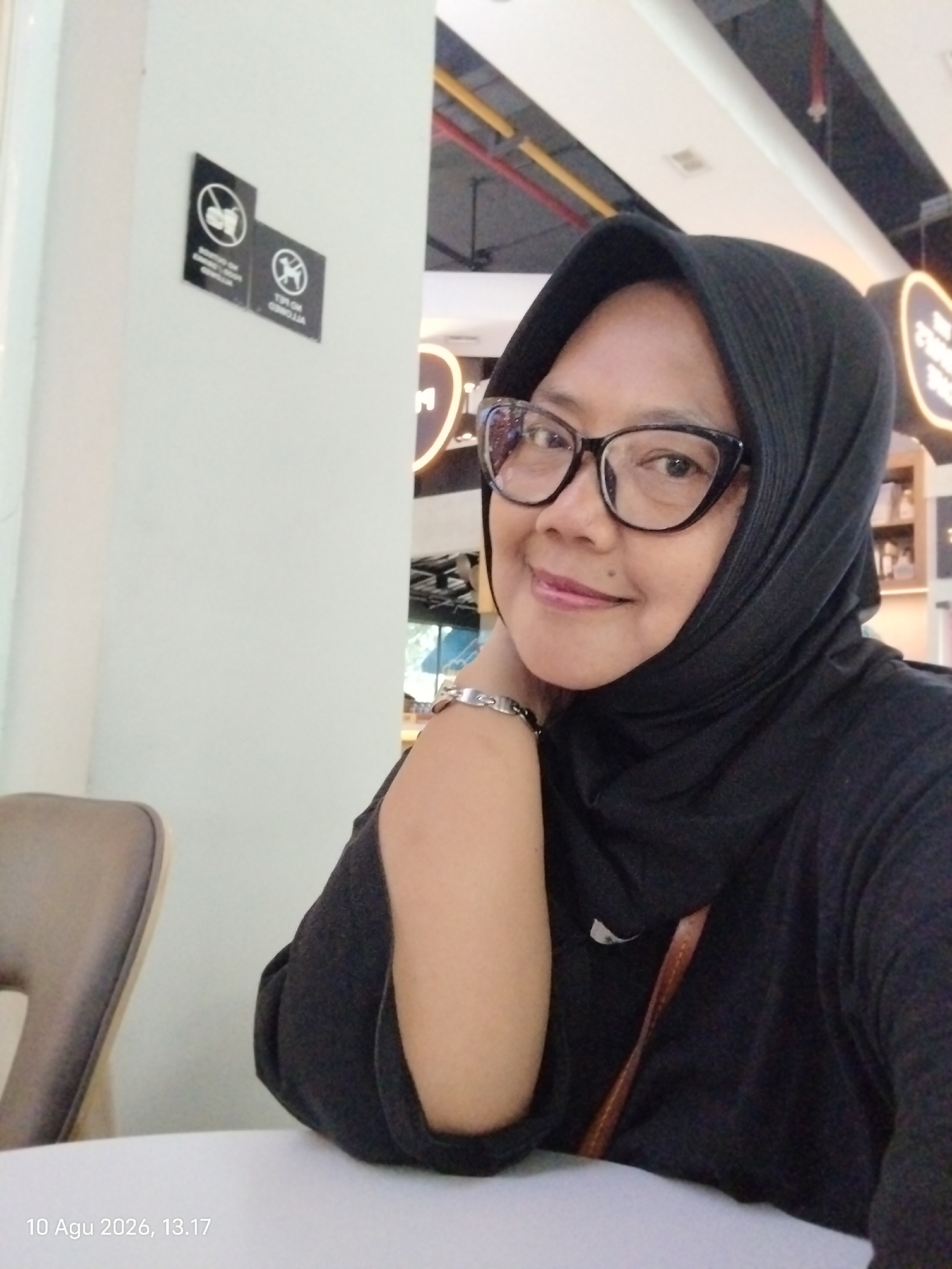 An Indonesian woman wearing a black hijab and glasses is pictured in a casual selfie. She has her right arm resting on her neck and is smiling. She is wearing a black t-shirt and a brown strap from a bag is visible across her chest. In the background, there are signs on a white wall, one depicting a "no smoking" symbol and the other a "no pets" symbol. The environment appears to be indoors, possibly a public space or a cafe, with overhead lighting and what looks like a ceiling structure with yellow and red accents. A chair is partially visible on the left side of the image. The date stamp "10 Agu 2026, 13.17" is visible in the lower left corner, suggesting the photo was taken in East Jakarta, Indonesia.
