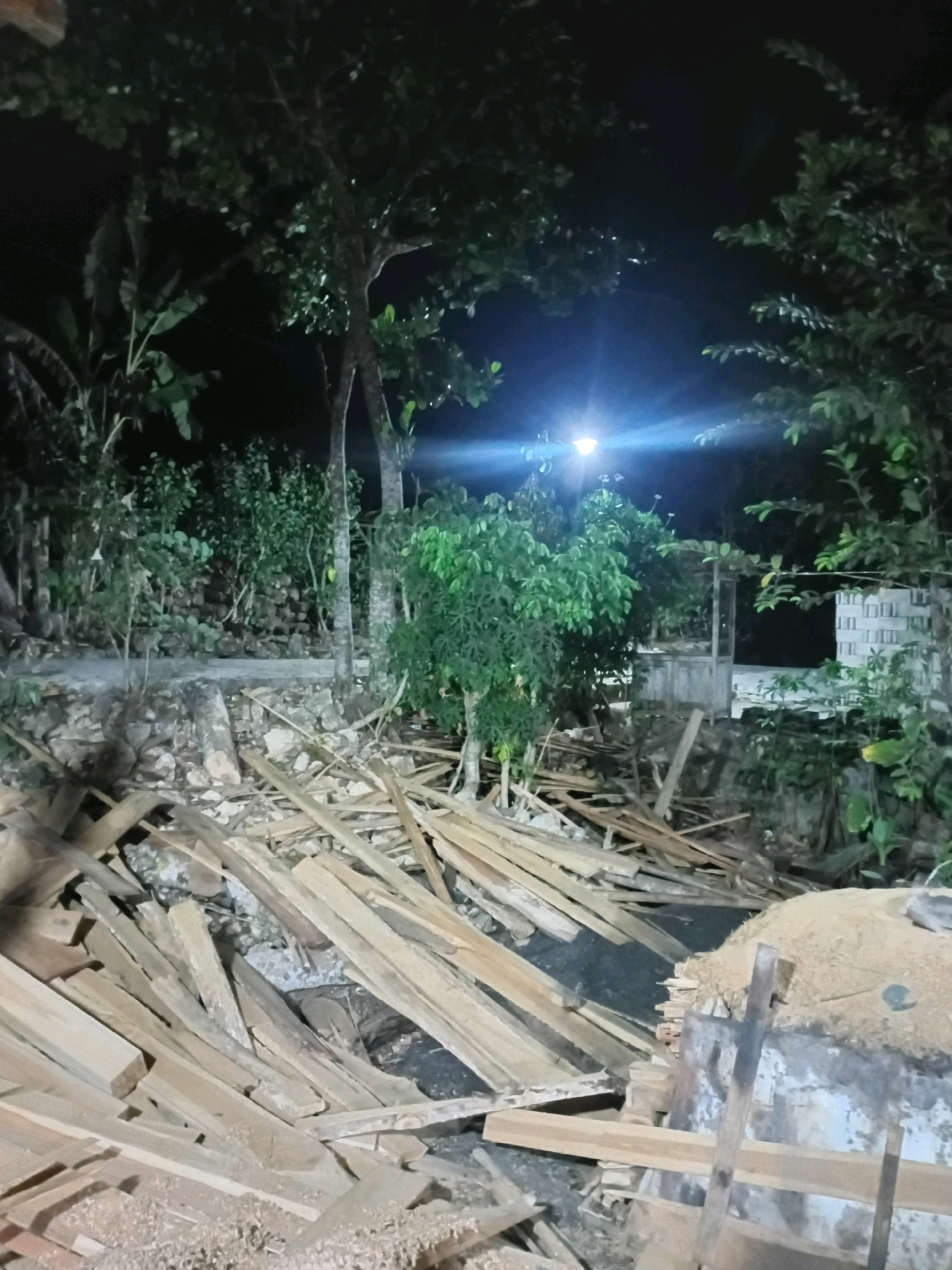 The image captures a nighttime scene in Playen, Indonesia, dominated by a large pile of cut lumber in the foreground, strewn across uneven ground. The lumber consists of various lengths and widths of unfinished wood, some appearing as planks and others as rougher pieces. Behind the lumber, the ground is a mix of dirt, rocks, and what appears to be a concrete structure. In the midground, a pile of granular material, possibly sawdust or sand, is visible next to a weathered, cylindrical object.
Further back, several trees with dense foliage are present, their leaves illuminated by a bright light source hanging from a pole in the upper center of the image, casting a noticeable glare. This light illuminates a path or walkway behind the trees. To the right of the trees, a white structure with horizontal rectangular elements is partially visible, suggesting a building. The overall environment appears to be outdoors, likely a yard or work area, with a sense of disarray due to the scattered lumber. The darkness of the night sky is contrasted by the artificial light, creating a scene that could be indicative of construction, demolition, or a lumber yard at night.