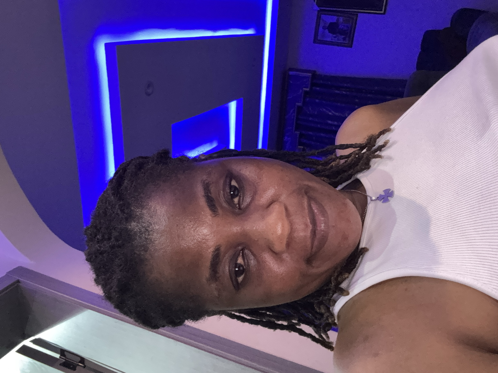 In a selfie taken indoors, a woman with dreadlocks and a white tank top smiles at the camera. She wears a pendant necklace. The background features a ceiling with blue neon lighting. The room appears to be a residential interior in Ajara, Nigeria.