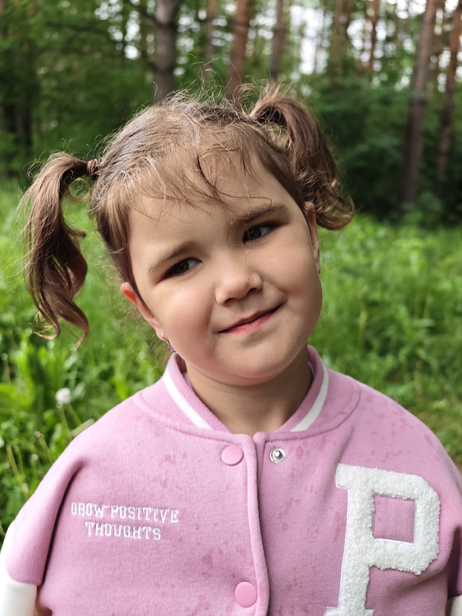 A close-up portrait shows a young child, likely a girl, outdoors in Chaikovsky, Russia. The child is positioned centrally in the frame, looking slightly to her right with a subtle, gentle smile. She has light brown hair styled in two pigtails, with some wispy strands around her face.
She is wearing a pink bomber-style jacket, featuring white stripes on the collar and cuffs. On the right side of the jacket, there is a large, white, fluffy letter "P". On the left side, white embroidered text reads "GROW POSITIVE THOUGHTS". The jacket is fastened with pink snap buttons.
The background is a soft blur of green foliage and tree trunks, suggesting a natural outdoor environment such as a forest or park. The lighting appears to be natural daylight, indicating it is daytime. The overall impression is one of calm and gentle innocence.