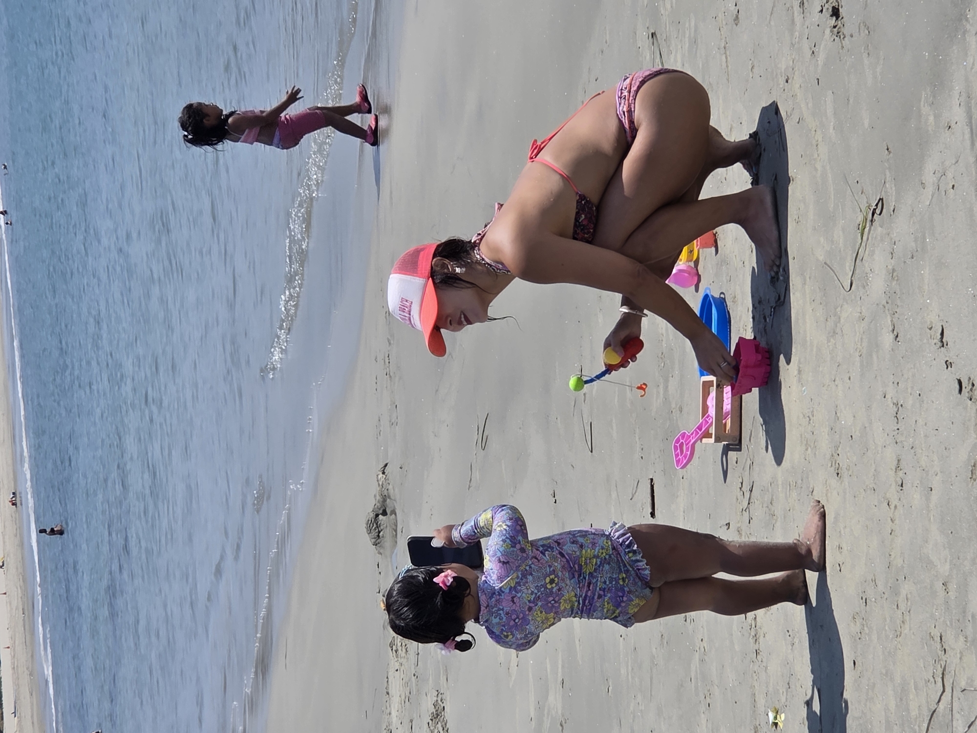 The image depicts a sunny day at the beach in Long Beach, USA, with three young girls engaging in activities. The scene is set on a sandy beach with the ocean in the background, suggesting a warm, possibly summer, day.
In the foreground, a girl in a colorful floral swimsuit is on her hands and knees, looking at a black object, possibly a phone. She has pink bows in her hair. To her right, another girl in a pink bikini and a red and white cap is also on her knees, interacting with beach toys. She is holding a yellow and red toy in her hand and there is a blue bucket and a pink shovel next to her. In the background, a third girl in a pink swimsuit is walking towards the ocean.
The overall mood seems playful and relaxed, indicative of a family outing at the beach. The lighting suggests it is daytime, likely mid-morning or afternoon, given the strong shadows. The sand appears damp near the water's edge, where small waves are breaking. Visible text on the girl's cap reads "LONG BEACH."