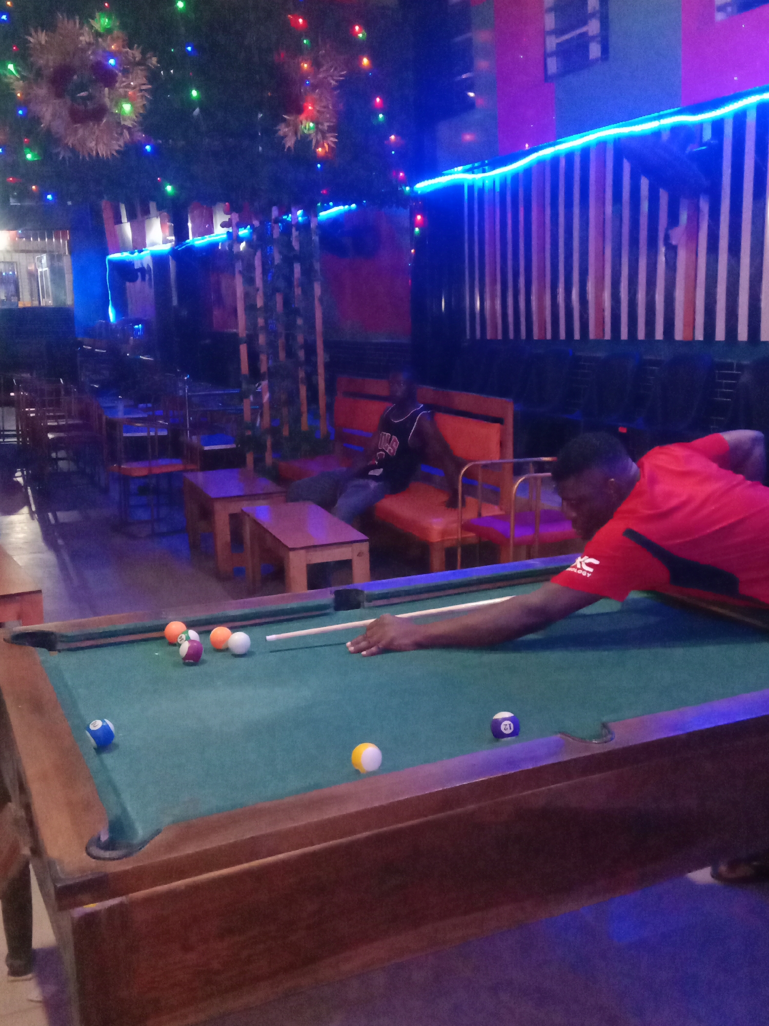 A man in a red shirt is playing pool in an establishment in Onitsha, Nigeria. He is leaning over the pool table, cue in hand, aiming at the cluster of balls. Another man is sitting on a bench in the background. The bar has tables and chairs, striped wall decor with blue lights, and festive decorations hanging from the ceiling.