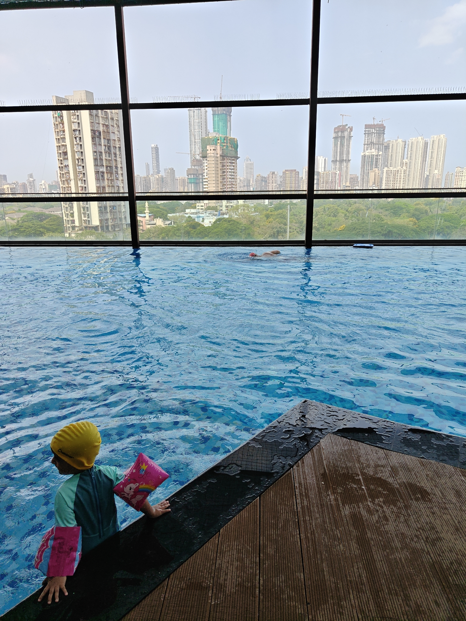 A slightly high-angle, medium shot shows a partially visible child standing in the shallow end of an indoor swimming pool, with an infinity edge overlooking the Mumbai, India, skyline.
The child is wearing a yellow swim cap, a long-sleeved aquamarine and blue swimsuit, and pink arm floaties. They are holding onto the edge of the pool with both hands, partially submerged in the water.
The pool is blue with visible wave patterns on the surface. The infinity edge of the pool is composed of black tiles. A dark brown, wood-slatted deck is partially visible in the foreground, adjacent to the pool edge.
Behind the pool is a large window framing a daytime cityscape of Mumbai, India. Tall buildings and construction sites are visible, with a green space in between the pool and the buildings. The city skyline is muted and partially obscured by the hazy atmosphere. Bird spikes are mounted on the exterior of the windows. Another person is seen swimming in the pool with a red cap.