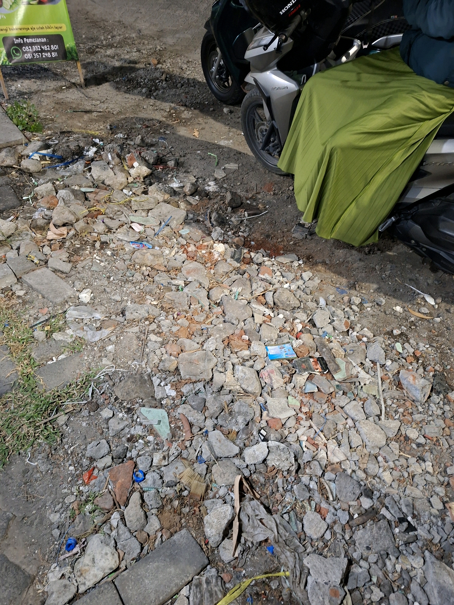 The image depicts a street scene in Surabaya, Indonesia. In the foreground, there is a pile of rubble and debris, including broken bricks, concrete fragments, and various pieces of trash. Some green grass is visible on the left side, growing amongst the debris. In the background, a motorcycle is parked. The motorcycle is silver and has a green cloth draped over its seat. Another motorcycle is partially visible on the right side. A green sign is also visible on the left, with text and a phone number displayed, suggesting a commercial establishment. The ground is uneven and appears to be unpaved or poorly maintained. The lighting suggests it is nighttime or dusk.