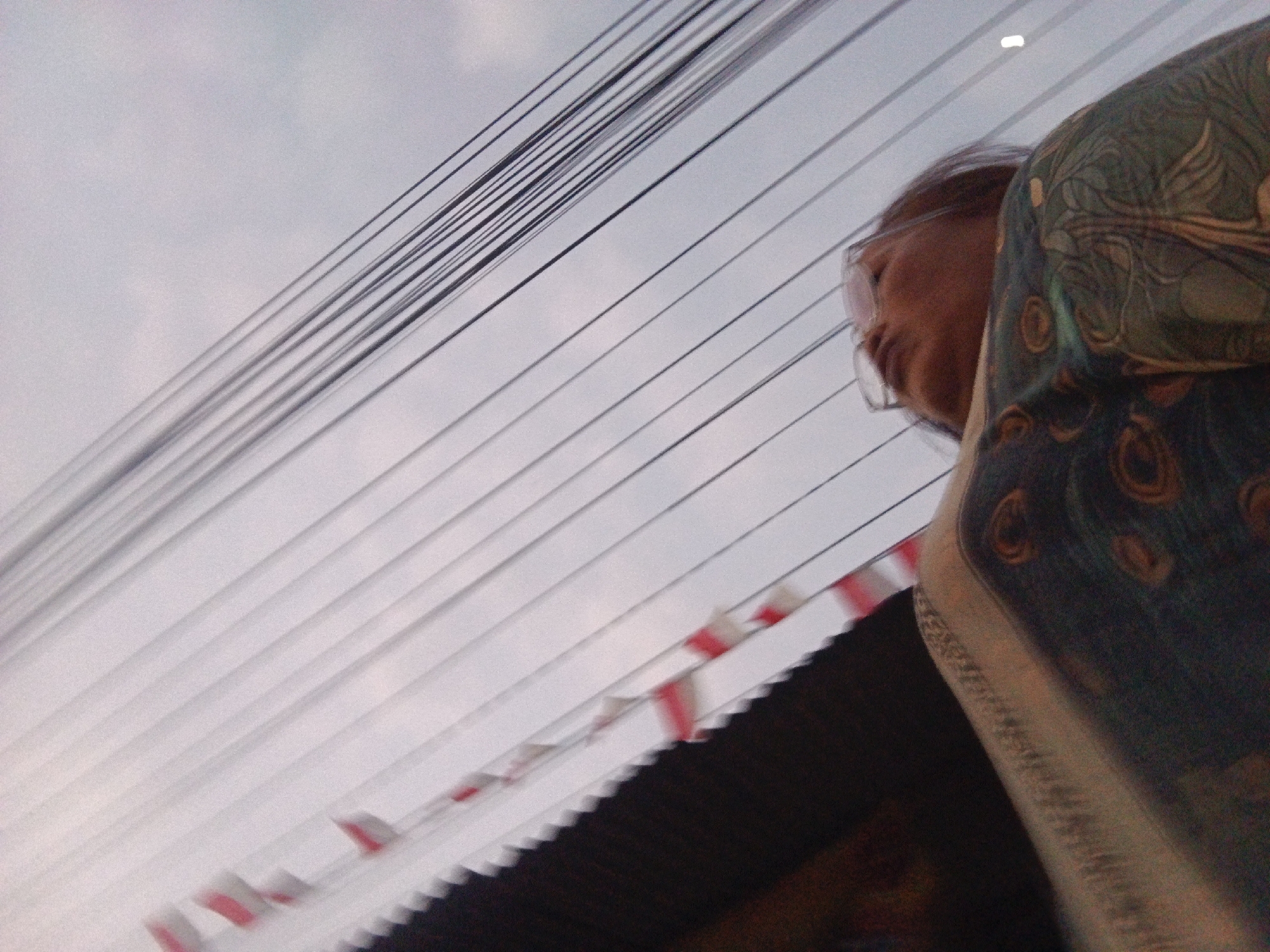 The image captures a low-angle perspective of overhead electrical wires against a pale sky, likely during daylight hours given the illumination. The wires are numerous and run diagonally from the upper left to the lower right of the frame. To the right, a portion of a person's head and shoulder is visible, wearing glasses and a garment with a dark green and teal peacock feather-like pattern. The person is looking downwards and to the left. In the lower portion of the image, a dark, textured surface is visible, possibly a roof or awning, with a decorative element consisting of alternating white and red rectangular shapes. The overall impression is a slightly blurred, candid shot. The image description does not provide any information about the specific location within Sukabumi, Indonesia, beyond what is generally visible in the photograph.