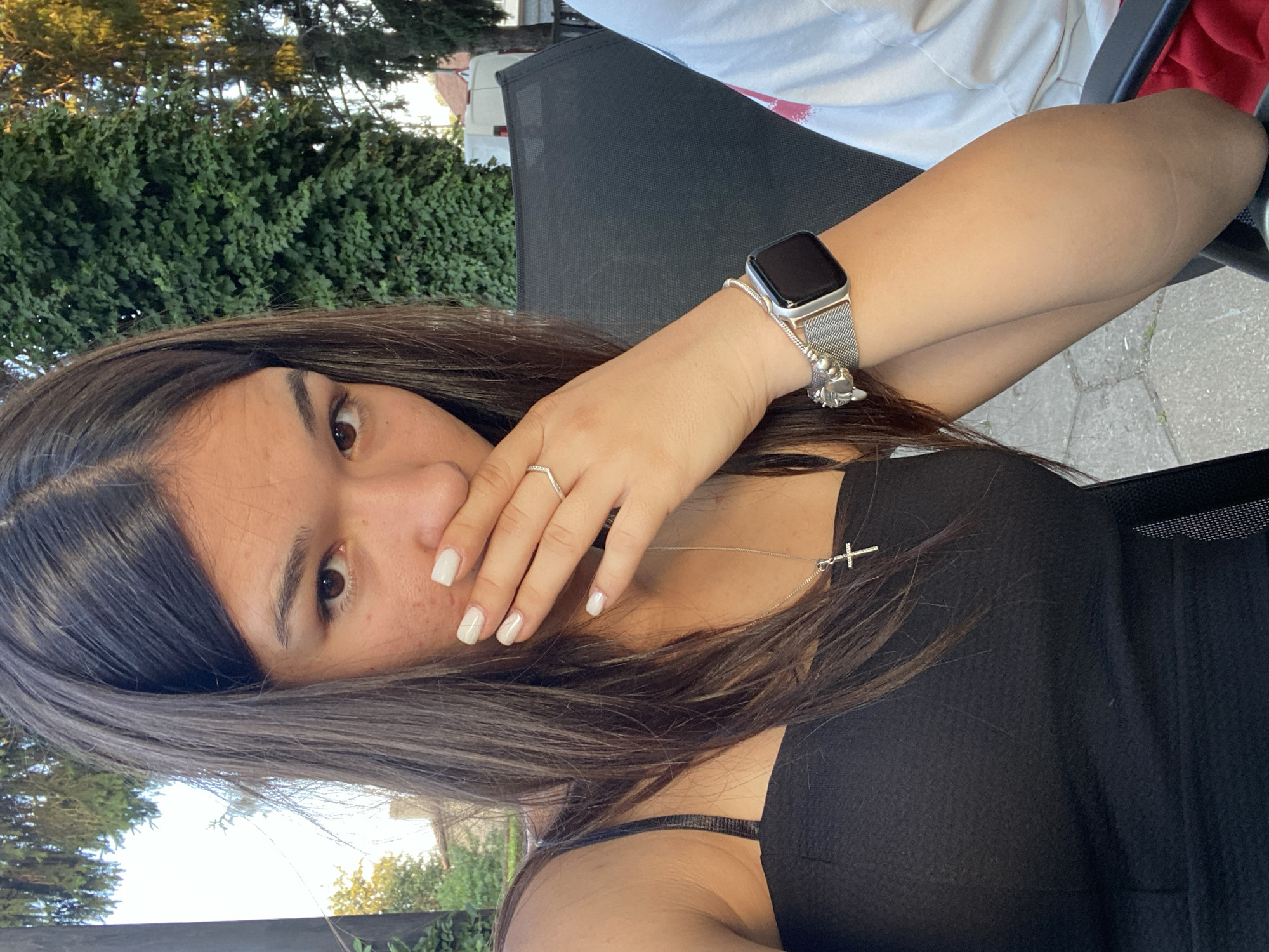 A young woman with long, dark hair is captured in a selfie, holding her hand over her mouth. She is wearing a black strapless top and a silver necklace with a cross pendant. On her left wrist, she has a silver smartwatch and a charm bracelet. The background appears to be an outdoor setting, with green foliage and trees visible. The woman is sitting in a black chair. The lighting suggests it is daytime, possibly late afternoon due to the warm glow on the trees. The environment suggests a relaxed, casual atmosphere. The exact location in Sevlievo, Bulgaria, cannot be determined from the visual information provided in the image alone.
