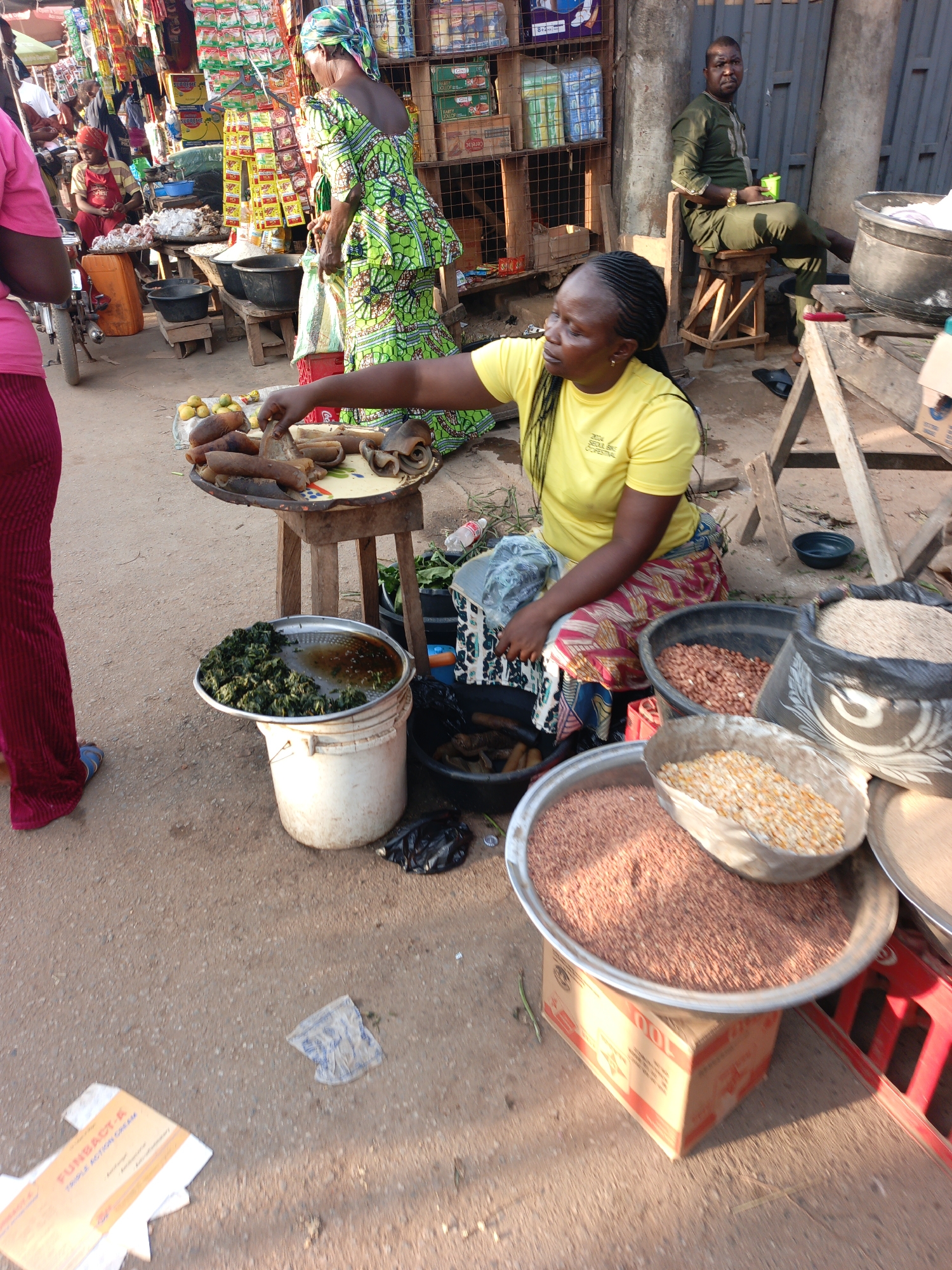 Akwanga, Nigeria. An outdoor market scene. A woman with braided hair wearing a yellow t-shirt and patterned skirt is seated next to several containers holding food items: cooked vegetables in water, peanuts, corn, and reddish-brown seeds. She reaches towards a tray of cooked meats on a small table. Another woman in a green patterned dress stands nearby. A man sits on a stool in the background. There is a building with a metal door, and several market stalls with various goods. The ground is dirt.