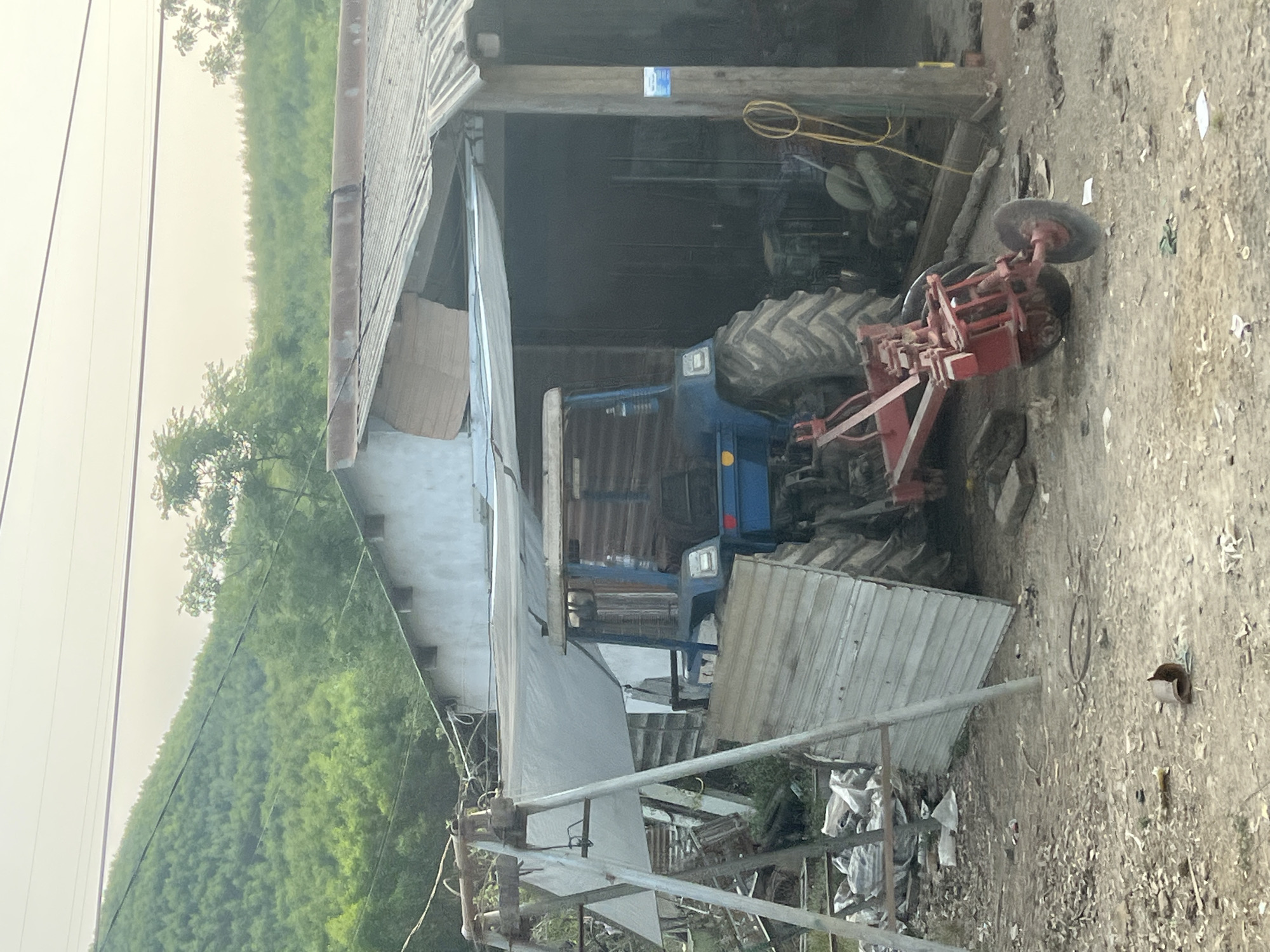 A blue tractor with a red attachment sits partially inside a shed structure. The shed has a corrugated metal roof and siding, with an open front. The surrounding environment is a dirt yard with scattered debris. In the background, a lush green hillside is visible. The photograph appears to have been taken near Myui, Vietnam.
