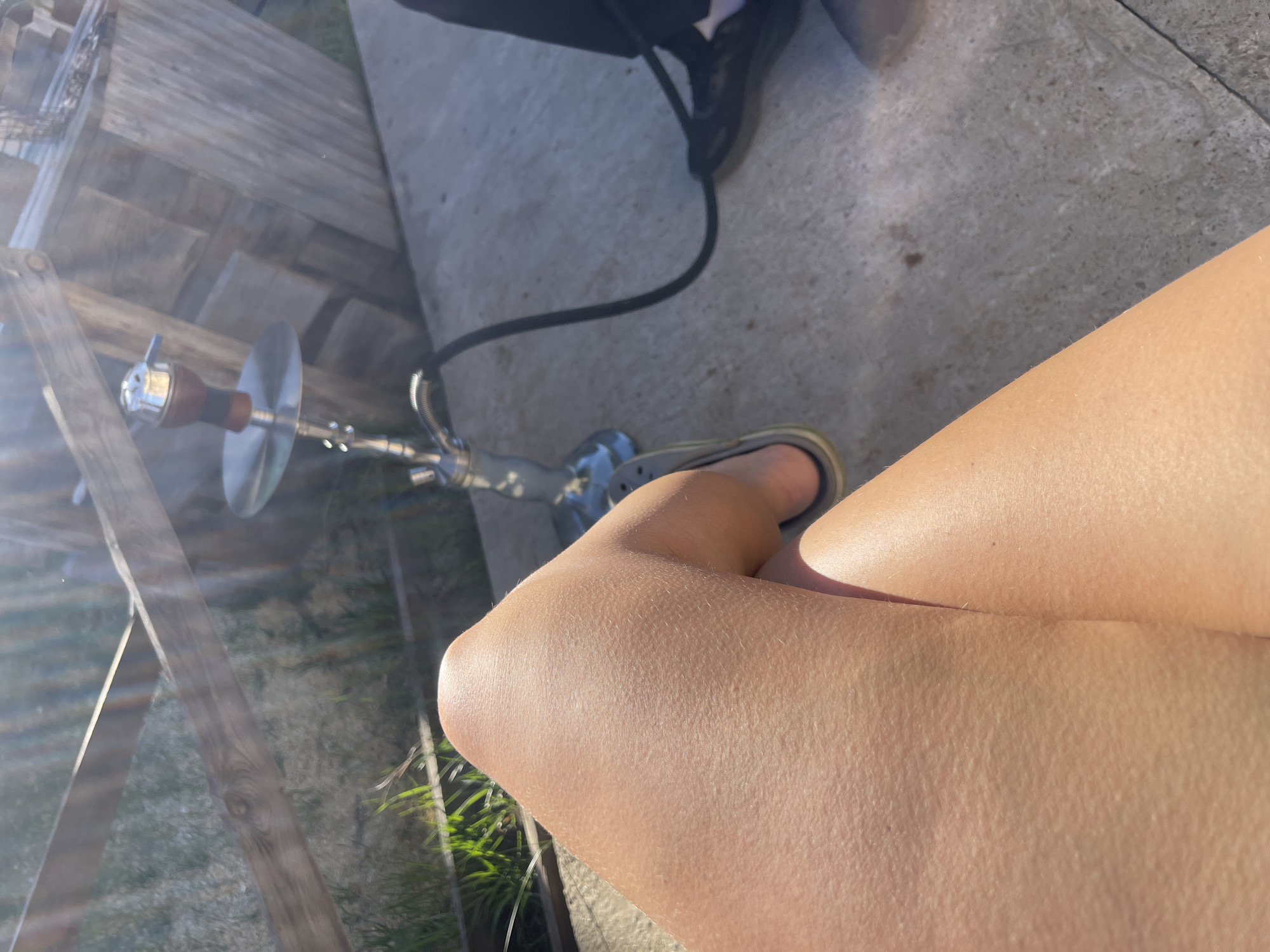 The image shows a portion of a person's crossed legs in the foreground, with a hookah placed on the ground. The legs are fair-skinned with visible hair follicles. One foot is visible, wearing a dark-colored sandal. A hookah sits on the ground, with a black hose extending to the left. The background includes concrete pavement, wooden boards, and sparse greenery. The location is unspecified, with no indications of it being Chaikovsky, Russia.