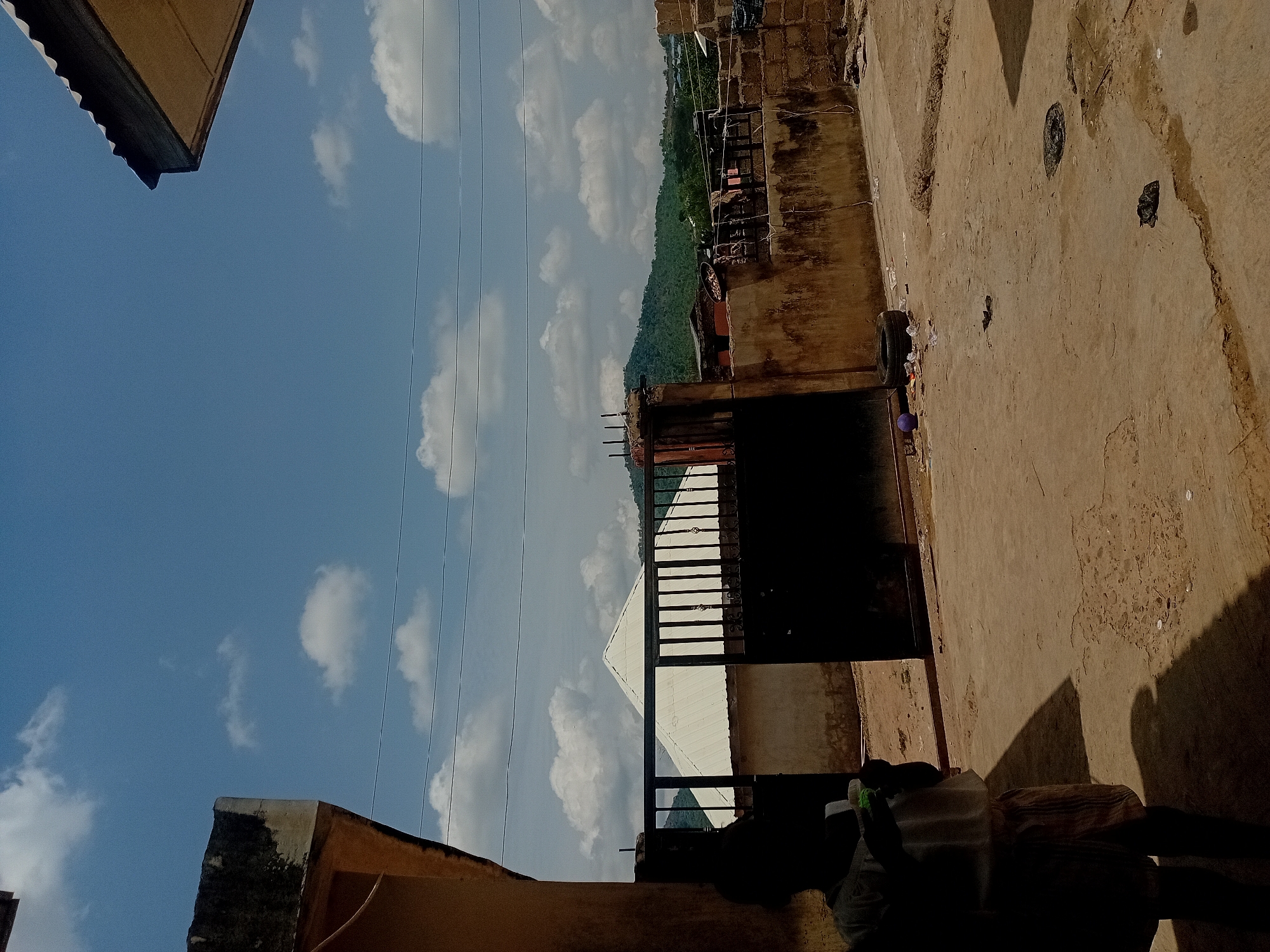 This image captures an outdoor scene from an elevated perspective, likely a rooftop or balcony, under a clear, sunny sky. The photo is rotated, presenting the sky vertically on the left side of the frame.
When oriented correctly, the upper half of the image features a bright blue sky adorned with scattered white cumulus clouds. Several thin, dark lines, resembling power lines or utility cables, traverse the sky horizontally. The top-left corner reveals the eave of a brown, corrugated roof structure, presumably part of the building from which the photo was taken.
In the middle ground, a dark metal gate or railing structure, possibly a balcony railing or part of a building entrance, is visible. Beyond this, a white building with a distinctive sloping roof stands out. Further in the distance, a prominent, verdant green hill or mountain range stretches across the landscape, suggesting a natural boundary to the urban or semi-urban environment.
The foreground, occupying the lower-right section of the frame, is a flat, textured, light brown surface, likely a concrete rooftop or yard. This surface exhibits signs of age and exposure, with darker patches and some scattered debris. Distinct, sharp shadows are cast upon this surface, indicating strong, direct sunlight. Among these shadows is a triangular shape and a dark, human-like silhouette, suggesting the presence of a person, possibly the photographer, just out of frame. A black tire and a small purple spherical object are also seen on this surface in the bottom-right corner. The bottom-left of the image shows a weathered, textured section of another building or wall.
The bright sky and pronounced shadows indicate that the photo was taken during the day, likely mid-morning or afternoon, under clear weather conditions. No identifiable text is visible within the image. The scene offers a view of both constructed elements and a natural landscape, hinting at a location where human habitation meets nature.