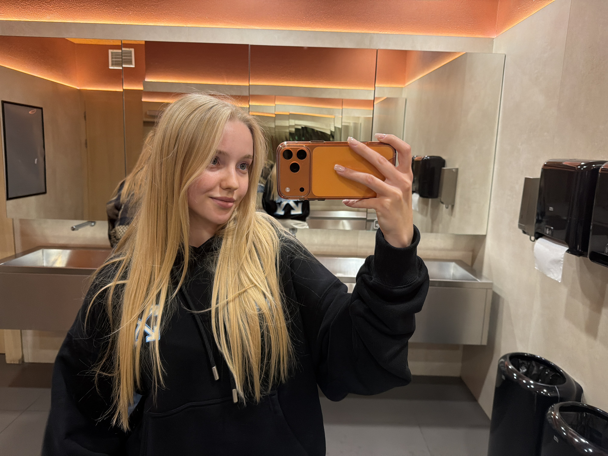 A young, fair-skinned woman with long blonde hair is taking a selfie in a public restroom. She is wearing a black hoodie with a logo visible on the left breast. Her left arm is visible, and she is holding an orange smartphone in her right hand to take the photo. The restroom features multiple stainless steel sinks and mirrors. There are black dispensers and trash cans mounted on the beige-colored walls. The lighting in the restroom is warm, with orange accents illuminating the space above the mirrors. The location is in the city of Unknown, Unknown.