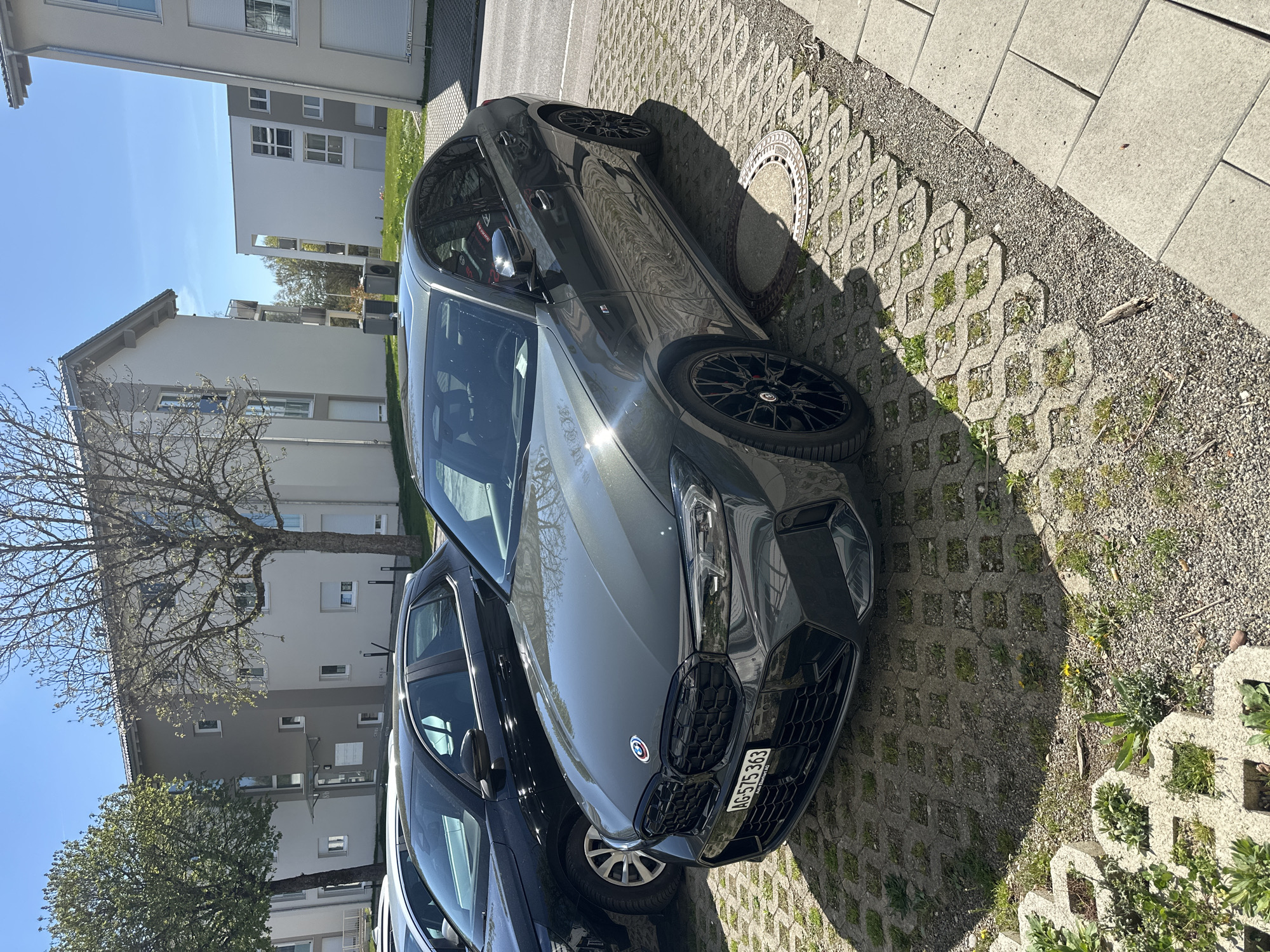 The image shows a gray BMW parked on interlocking concrete pavers in a residential area of Jestetten, Germany. A black vehicle is partially visible to the left of the BMW. Buildings and trees are in the background under a blue sky. The license plate of the BMW reads "AG-575363". A manhole cover is near the front tire of the BMW.