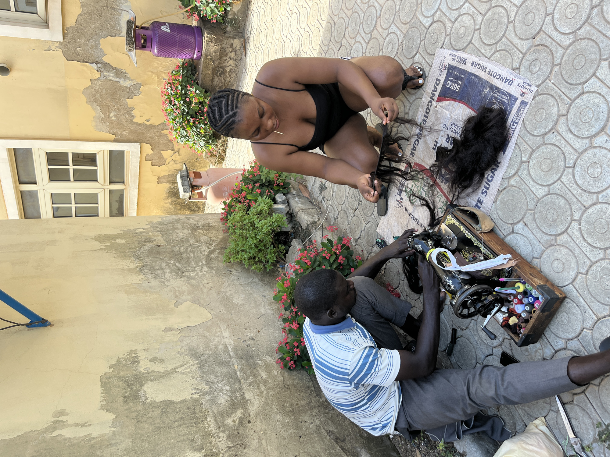 The image captures an outdoor scene in what appears to be a residential area in Abuja, Nigeria. A woman wearing a black top is kneeling on a patterned pavement, seemingly working with strands of dark hair. Beside her lies a white sack labeled "DANGOTE SUGAR 50KG." A man in a blue and white striped shirt and grey pants is sitting on the pavement, operating a black sewing machine. Next to him is a wooden box containing colorful spools of thread and other sewing accessories. In the background, a yellow building with peeling paint can be seen, along with windows and pink flowering plants. A purple gas cylinder is also visible in the background.