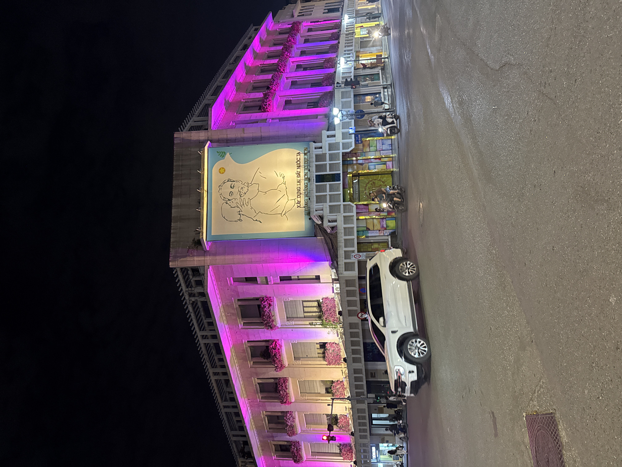 The image depicts a nighttime street scene in Hanoi, Vietnam. A large, multi-story building is illuminated with pink and purple lights, featuring a mural of a man and child on its facade. Flower boxes adorn the windows of the building. In the foreground, a white SUV is parked on the wet asphalt street, suggesting recent rain. Several motorcycles are also visible on the street, with riders. Traffic lights are present, and streetlights illuminate the area. The environment appears to be an urban setting with a mixture of commercial and residential elements.